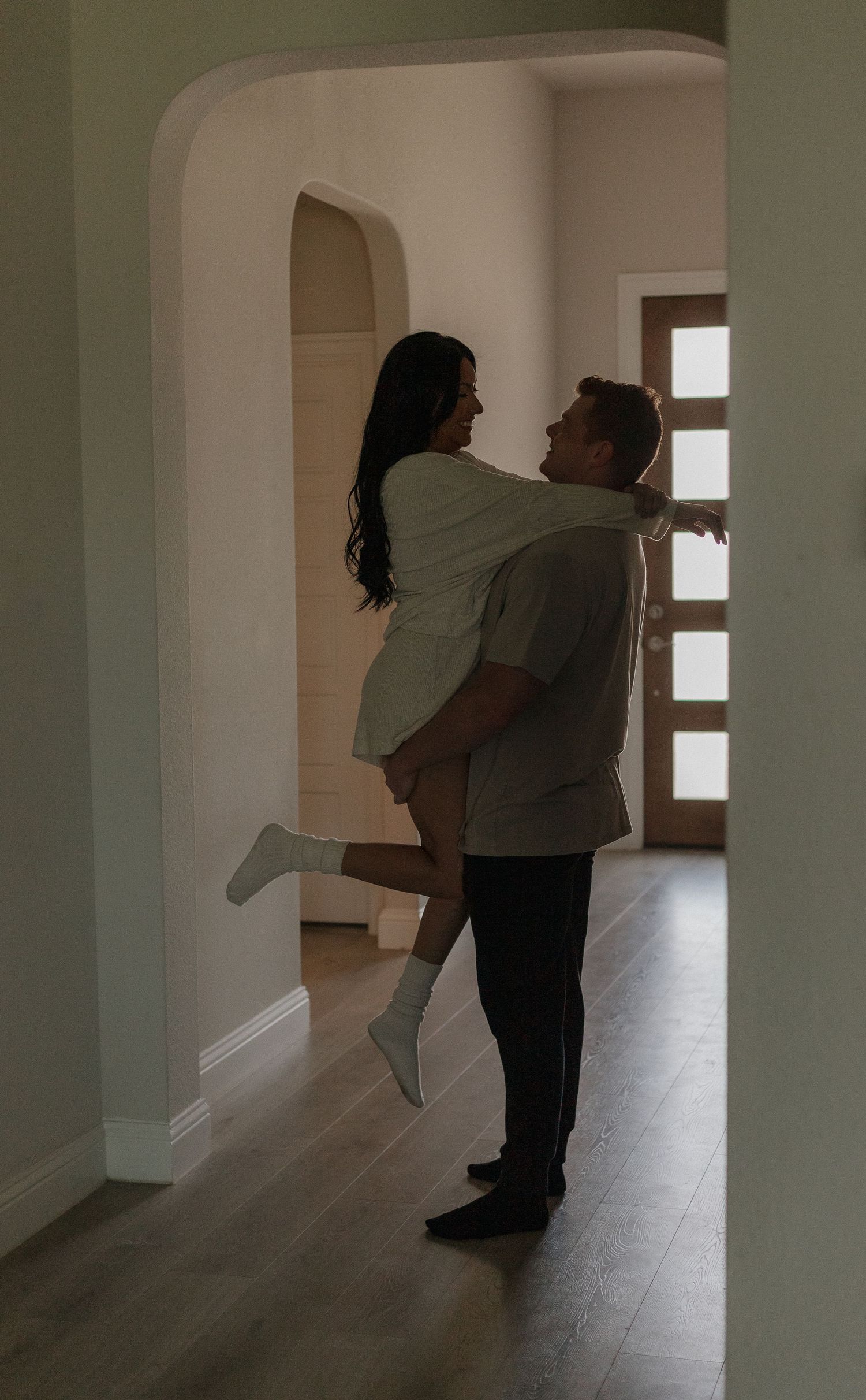 A romantic couple embraces in a hallway with arched doorways and hardwood floors, creating an intimate silhouette.