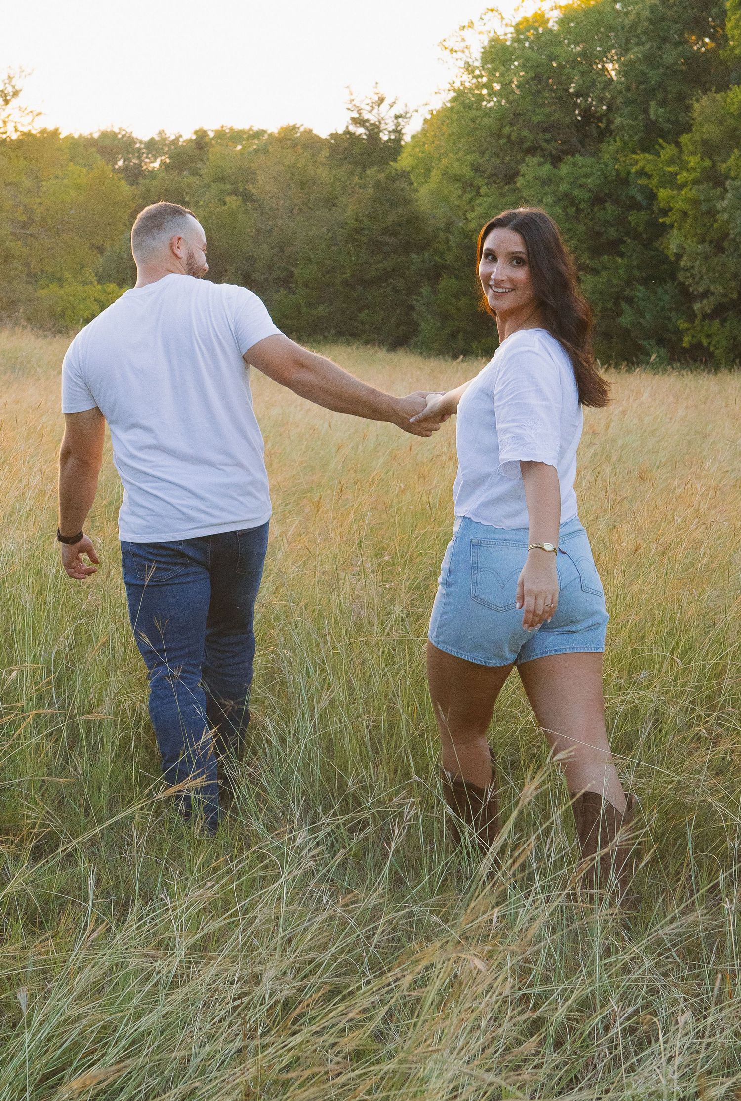 A couple walks hand in hand through tall grass at sunset.