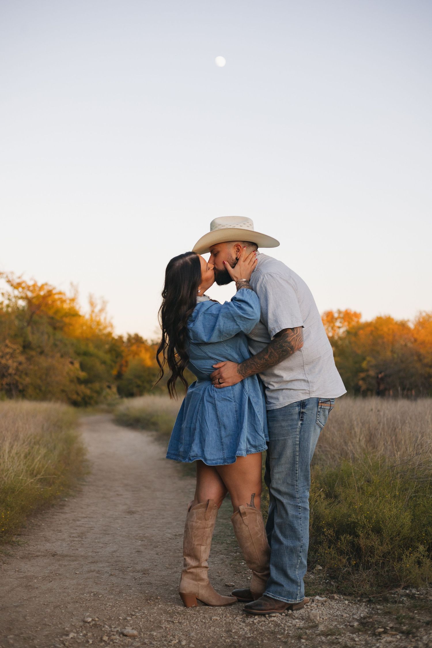 A couple in denim and western wear share intimate moments along a rustic dirt path at sunset.