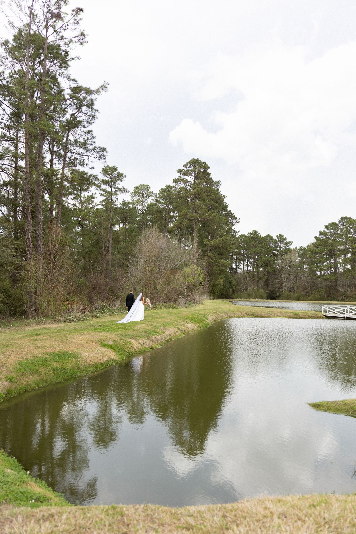 A scenic pond with tall pine trees reflects the cloudy sky while a couple takes wedding photos along the grassy bank.