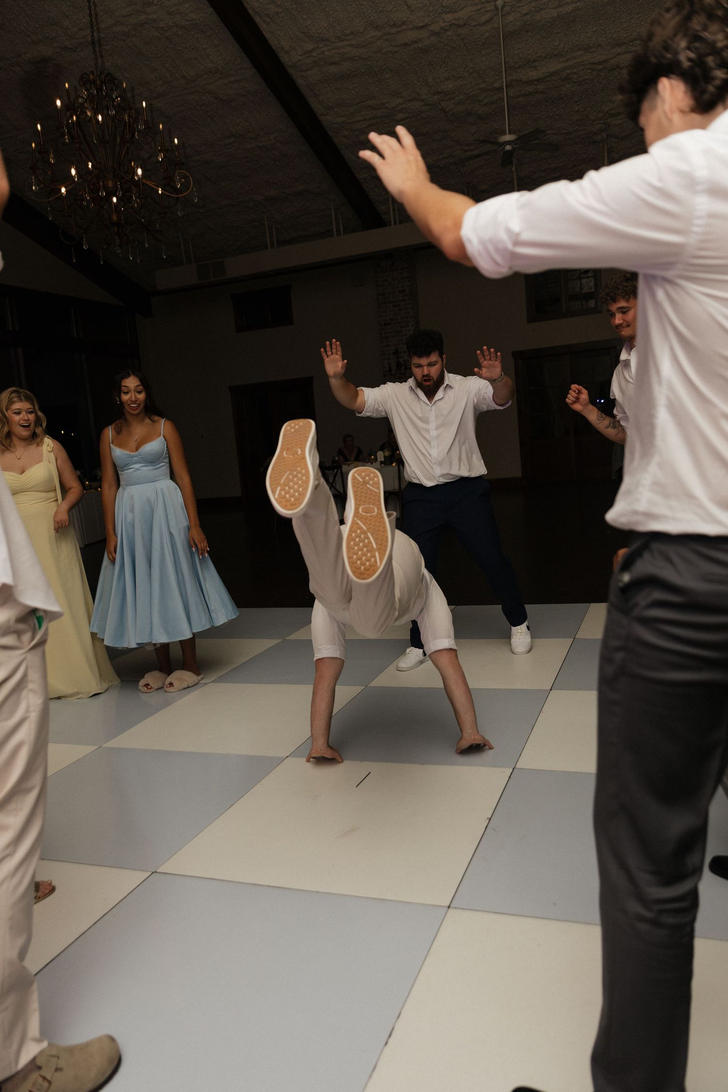 A guest breakdances on a checkered dance floor while wedding guests watch and cheer at a reception.