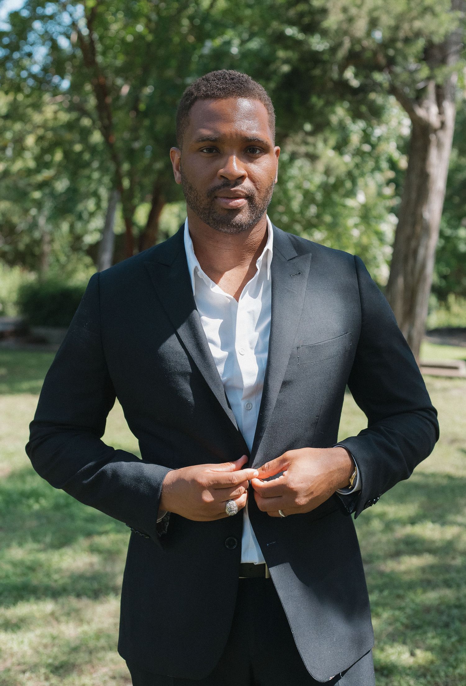 Groom in black suit adjusts his jacket buttons in outdoor garden setting.