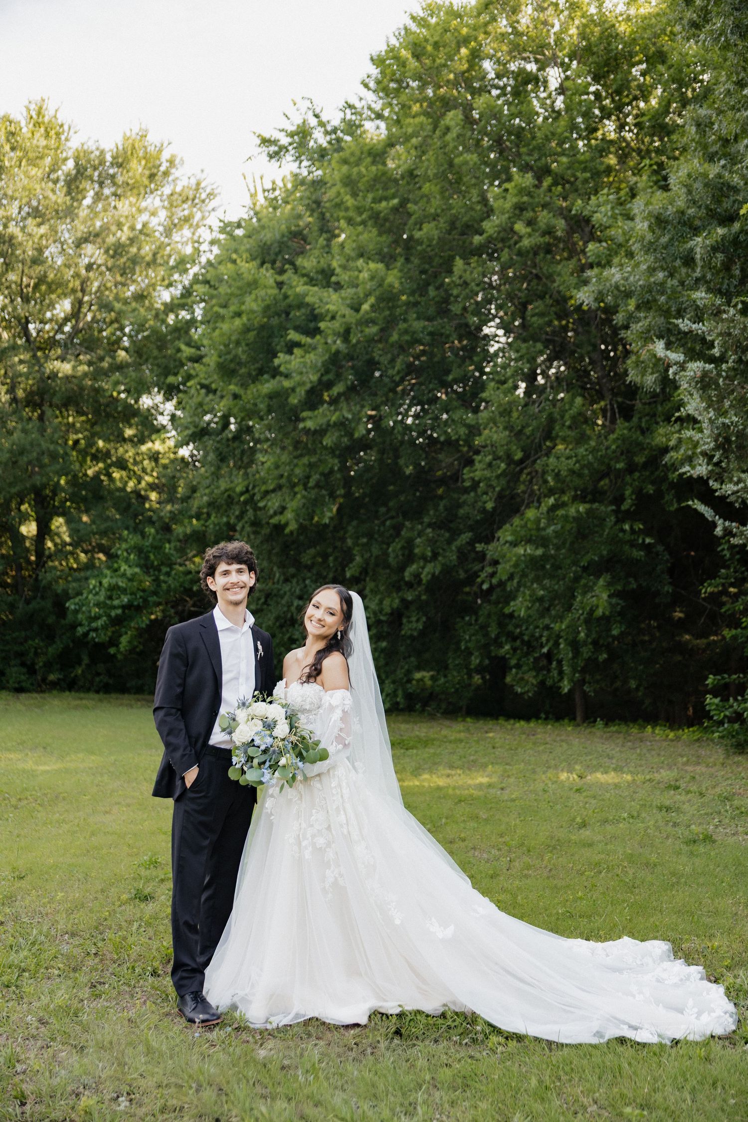 A bride and groom pose outdoors on green grass with lush trees, she wears a floral gown with long veil.