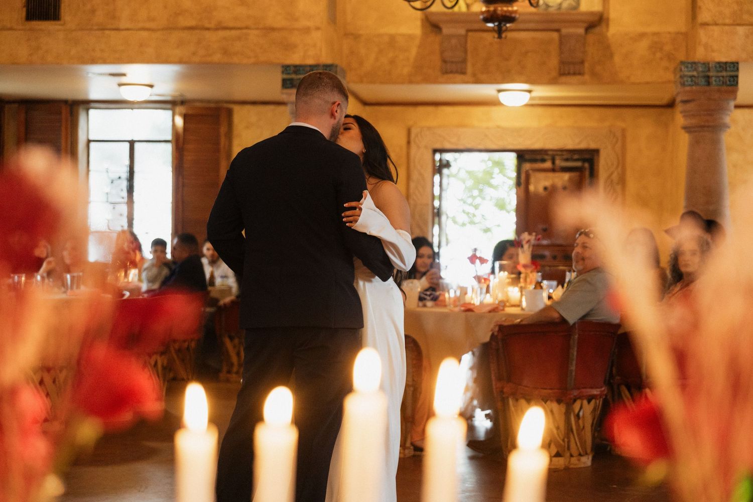 Couple shares first dance at elegant wedding reception with glowing candles and red floral arrangements in warm candlelit venue.