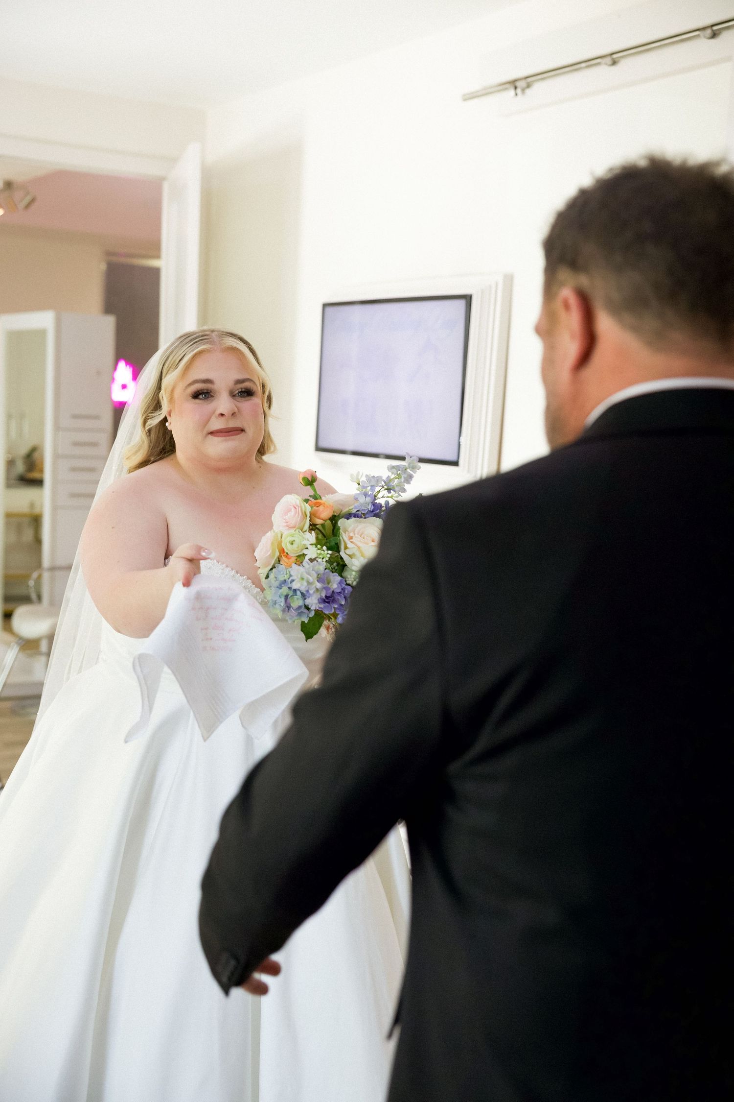 Bride in white gown holding colorful bouquet during emotional first look moment with groom indoors.