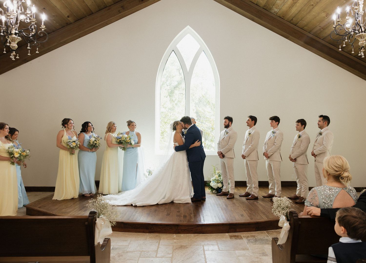 Bride and groom share first kiss at chapel altar surrounded by bridesmaids in pastel gowns and groomsmen in tan suits.
