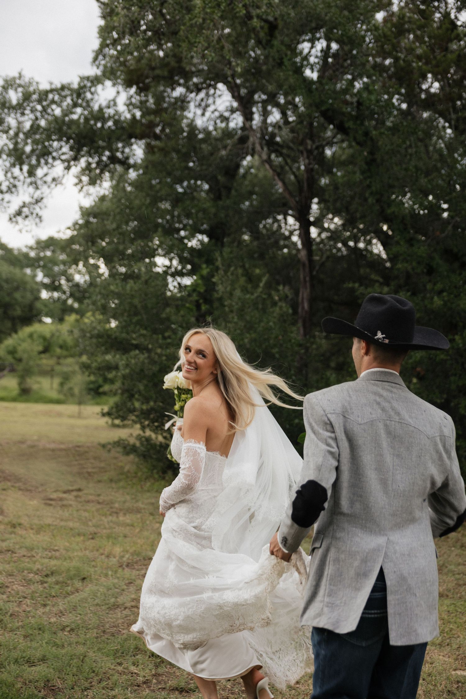 A smiling bride in a white gown holds flowers while walking hand-in-hand with a groom in a cowboy hat outdoors.