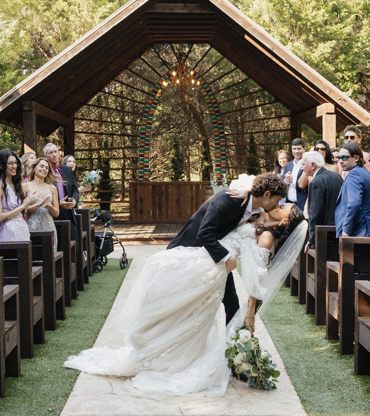 Bride and groom share a romantic dip kiss at outdoor woodland wedding chapel aisle, surrounded by cheering guests.