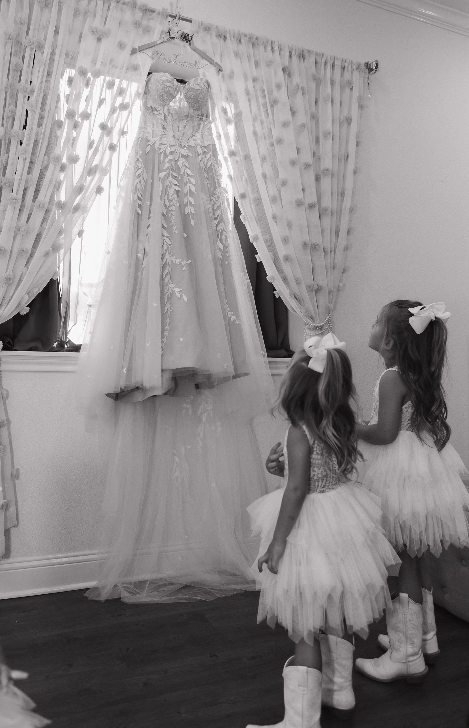 Two flower girls in white dresses admire a hanging wedding gown against a window with polka dot curtains.