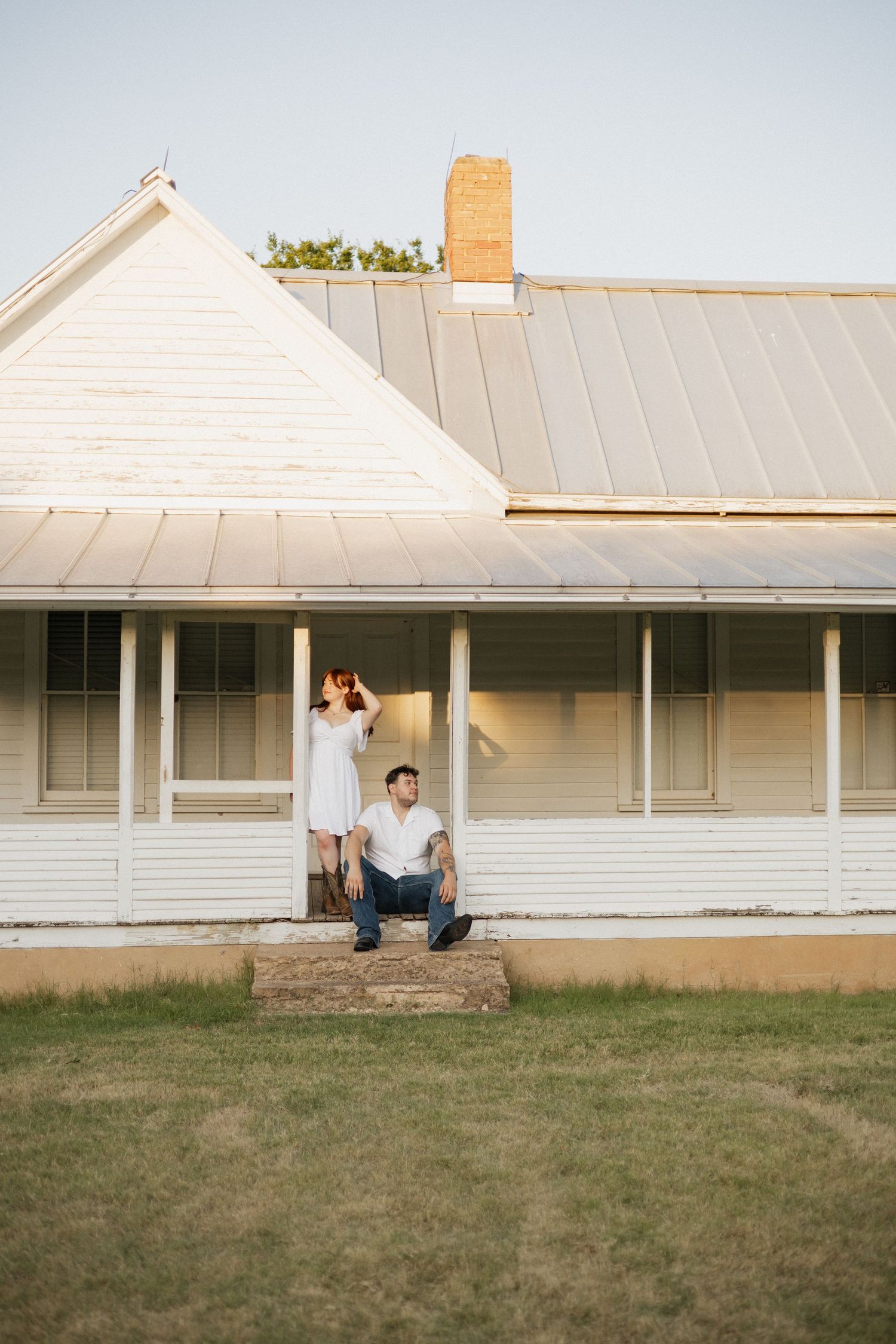 Couple posing in front of a white farmhouse with metal roof, man sitting on steps, woman standing behind him.