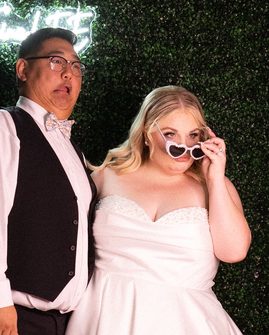Bride in white strapless gown with heart sunglasses poses with groom in vest and bow tie at wedding photo booth.