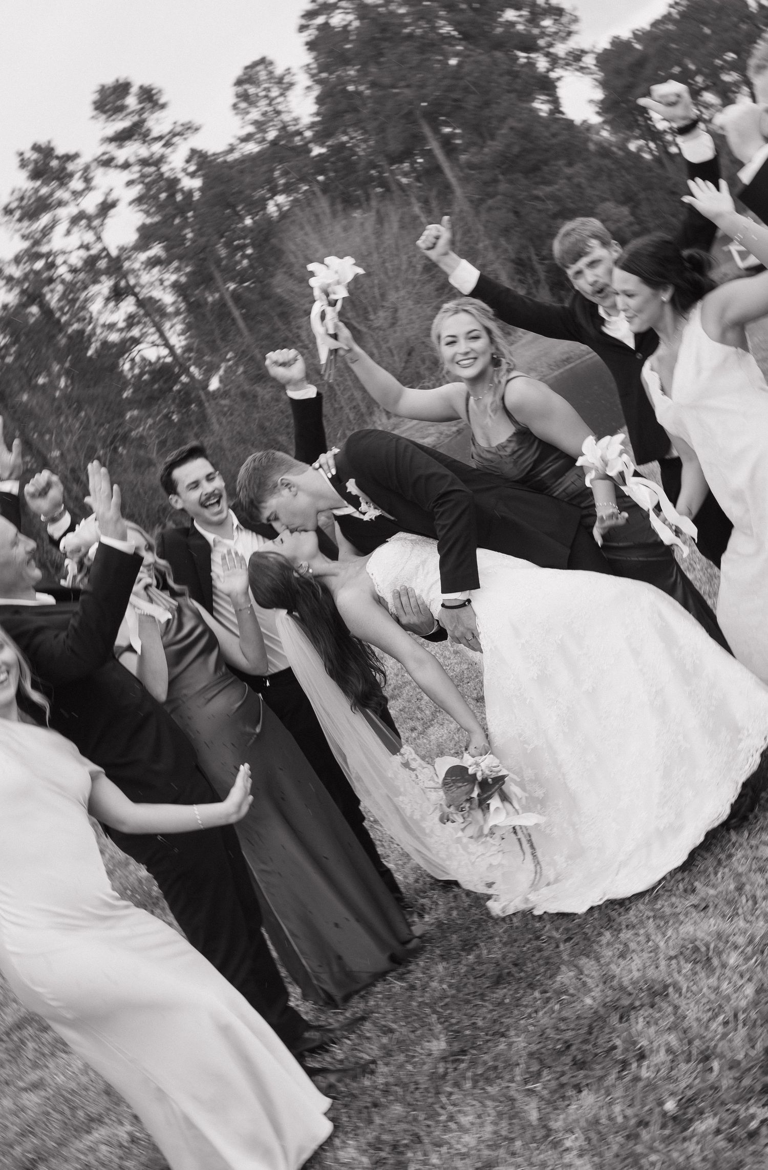 A joyful black and white photo of a wedding party jumping and celebrating outdoors with flowers in their hands.