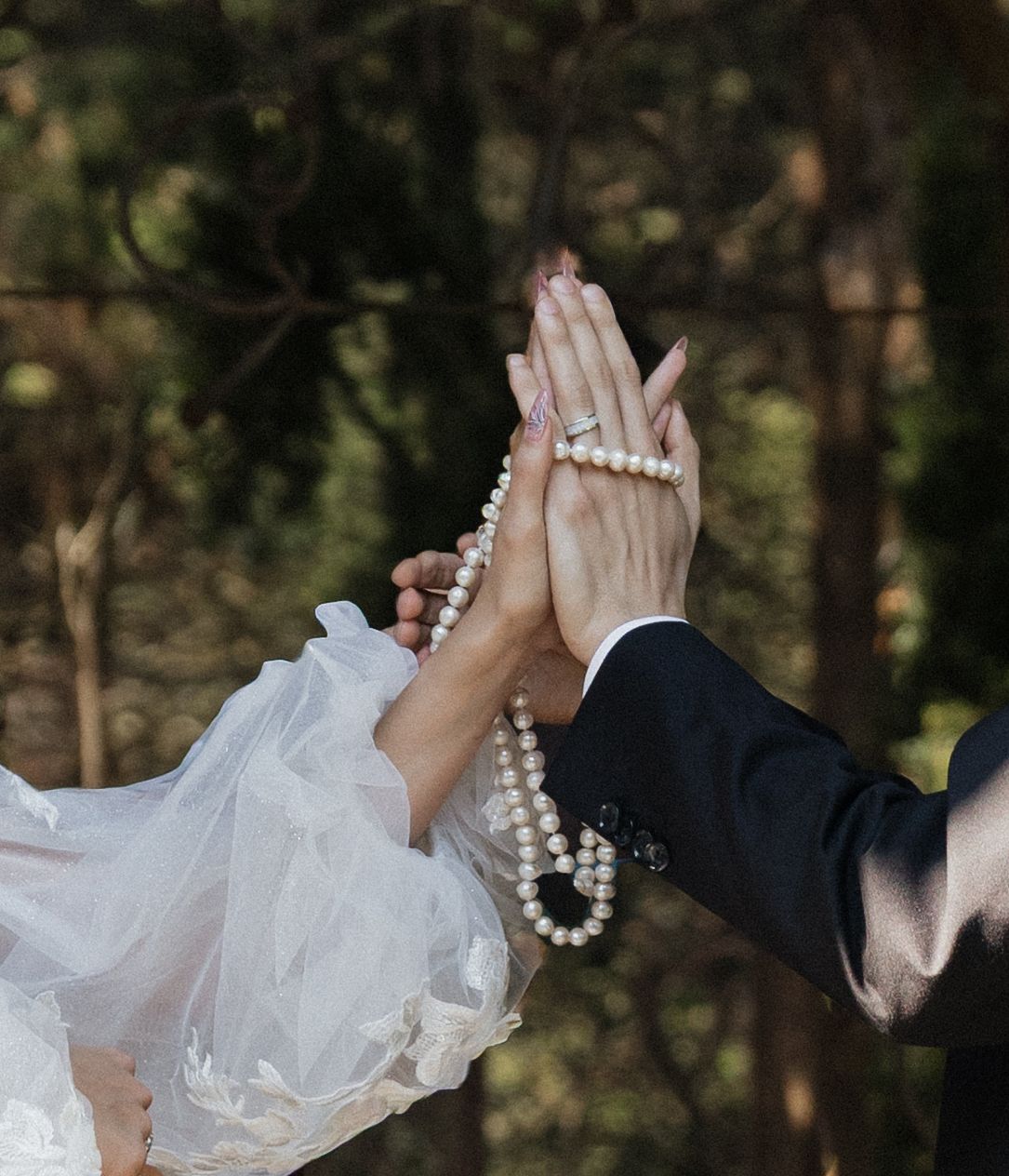 Bride and groom joining hands with pearl necklace during outdoor wedding ceremony in forest setting.