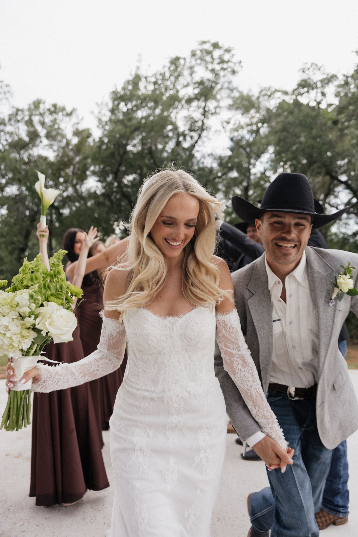 Smiling bride in lace off-shoulder gown holds bouquet while groom in cowboy hat and jeans walks hand-in-hand after ceremony.