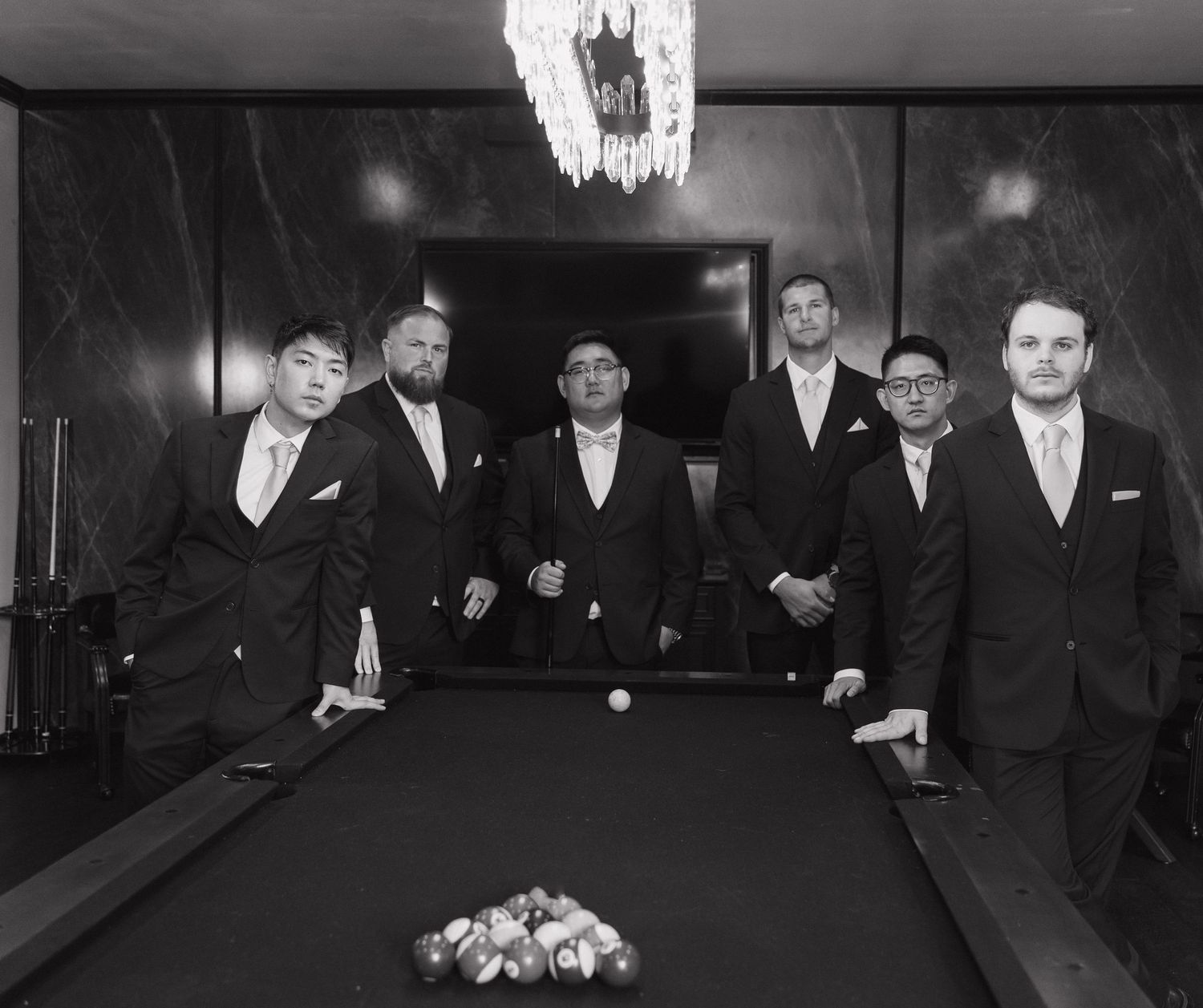 Group of six groomsmen in dark suits posing around a billiard table in a sophisticated black and white photo.
