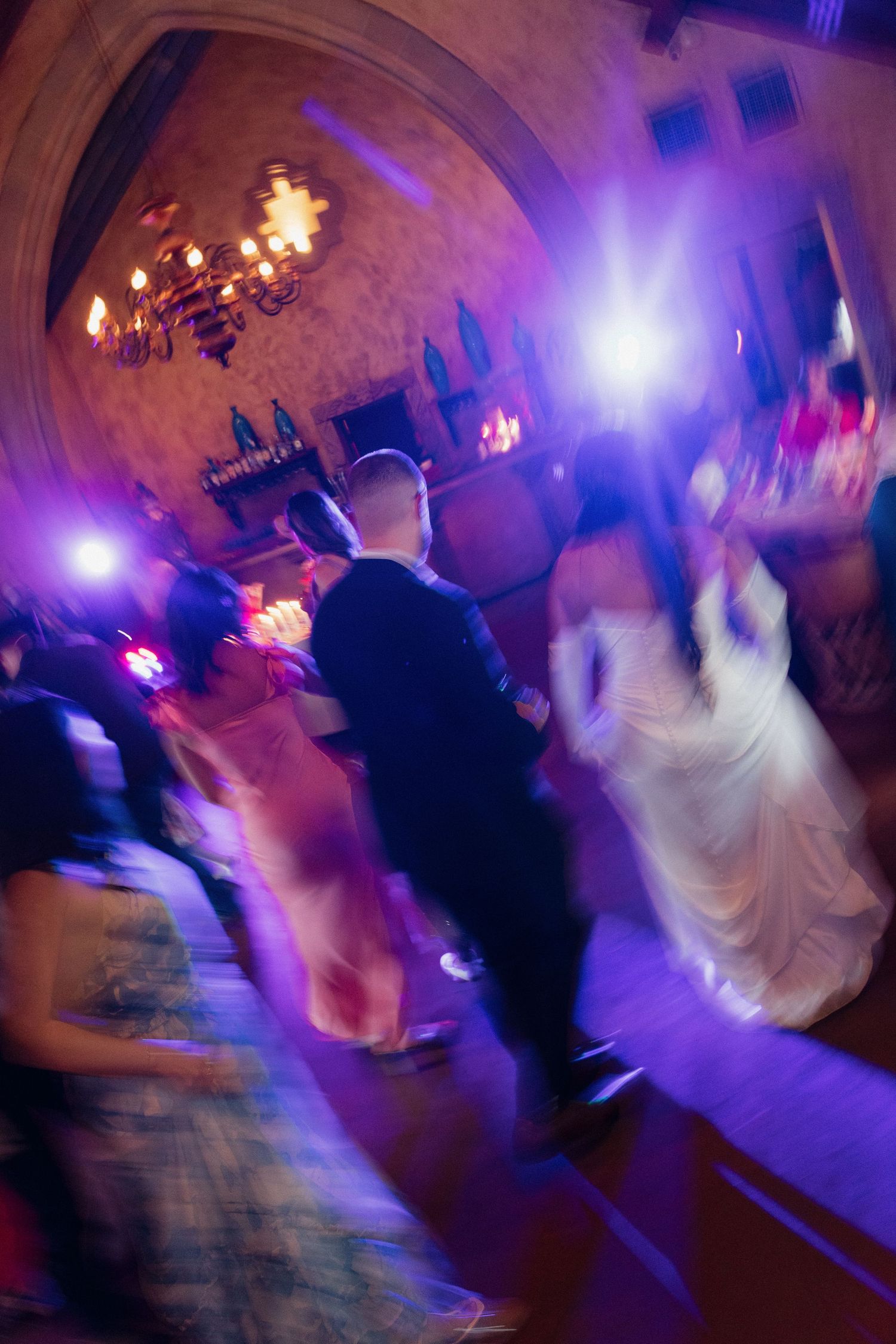 Bride and groom dance with guests under colorful lights at wedding reception.