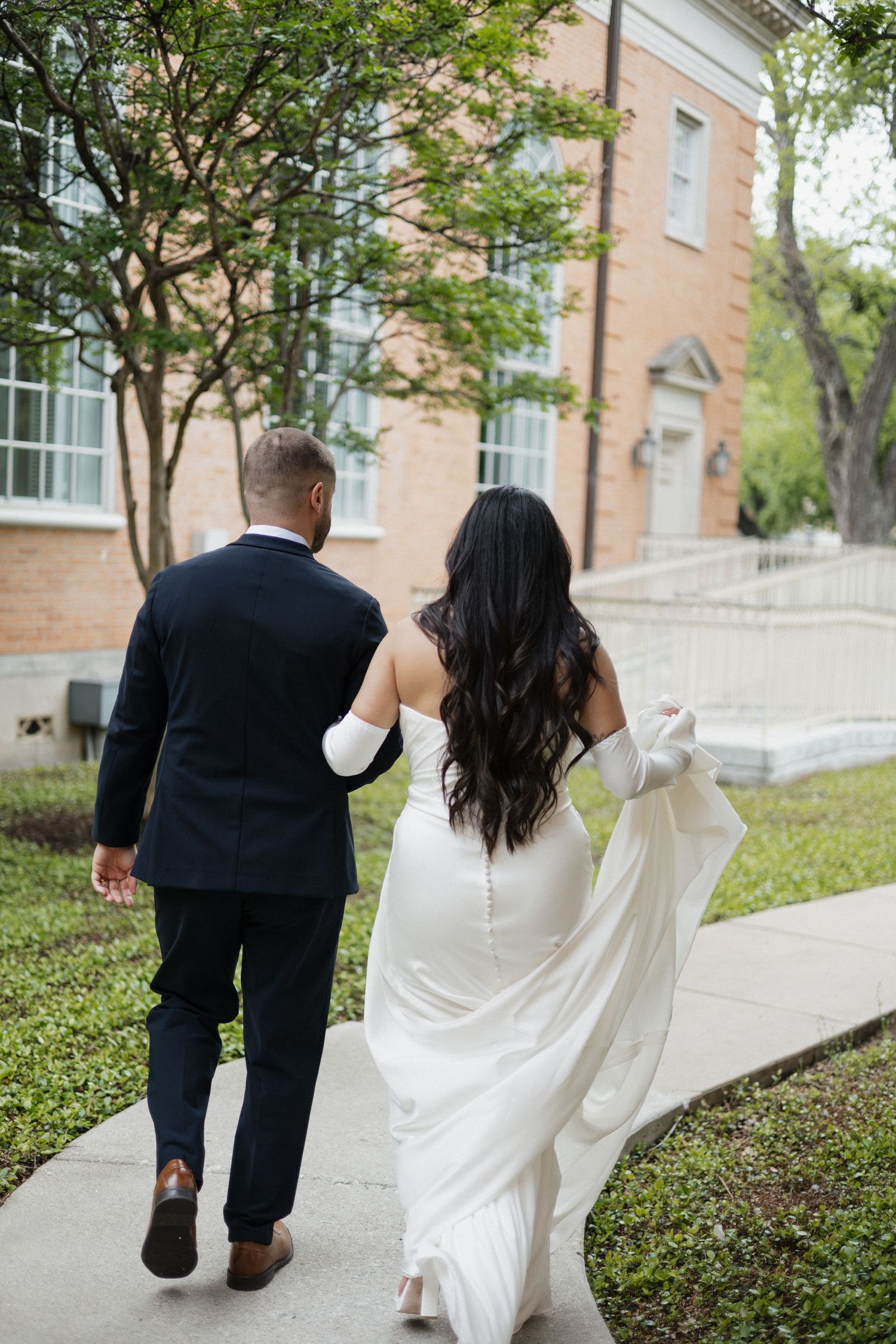 Bride and groom walking hand in hand along a sidewalk outside a brick building on their wedding day.
