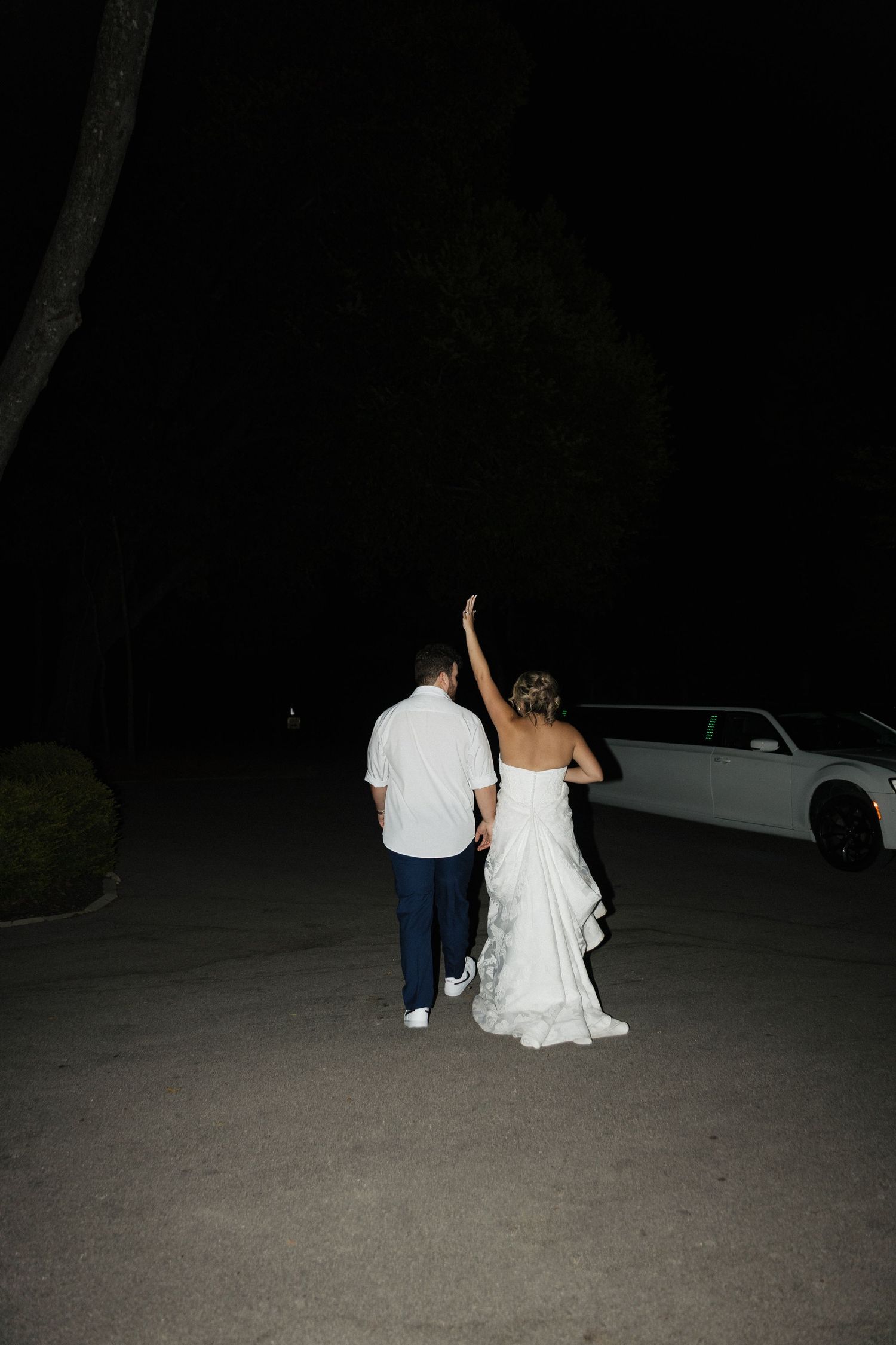 Newlywed couple walks hand-in-hand toward a white limousine at night, bride waving goodbye after wedding reception.