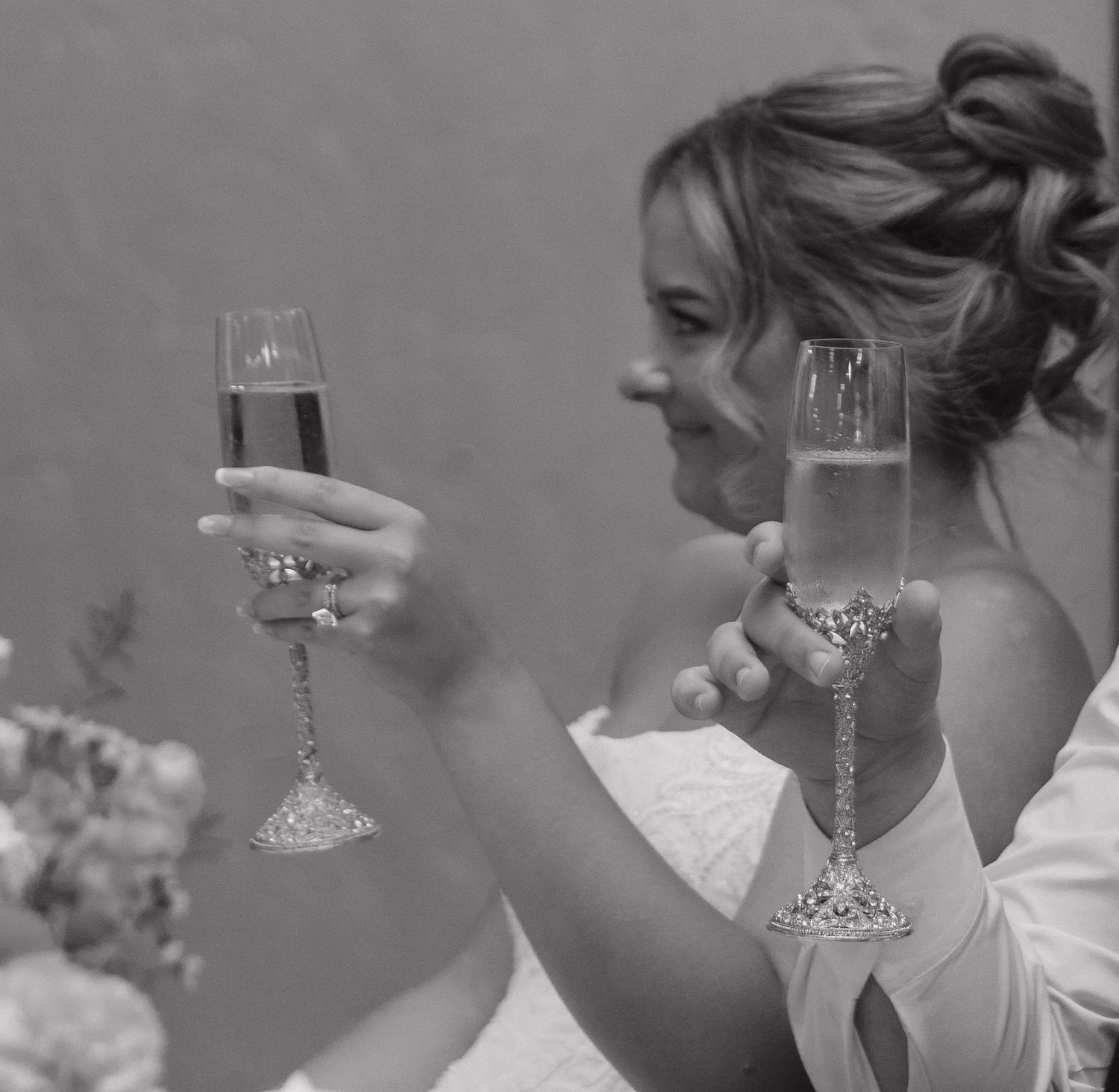Bride in white dress holding two decorative champagne flutes in a black and white wedding celebration photo.
