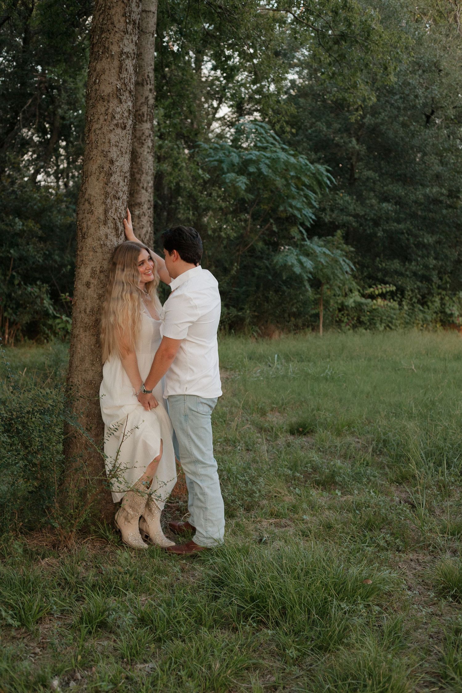 A romantic couple shares an intimate moment against a tree trunk in a serene outdoor setting at dusk.