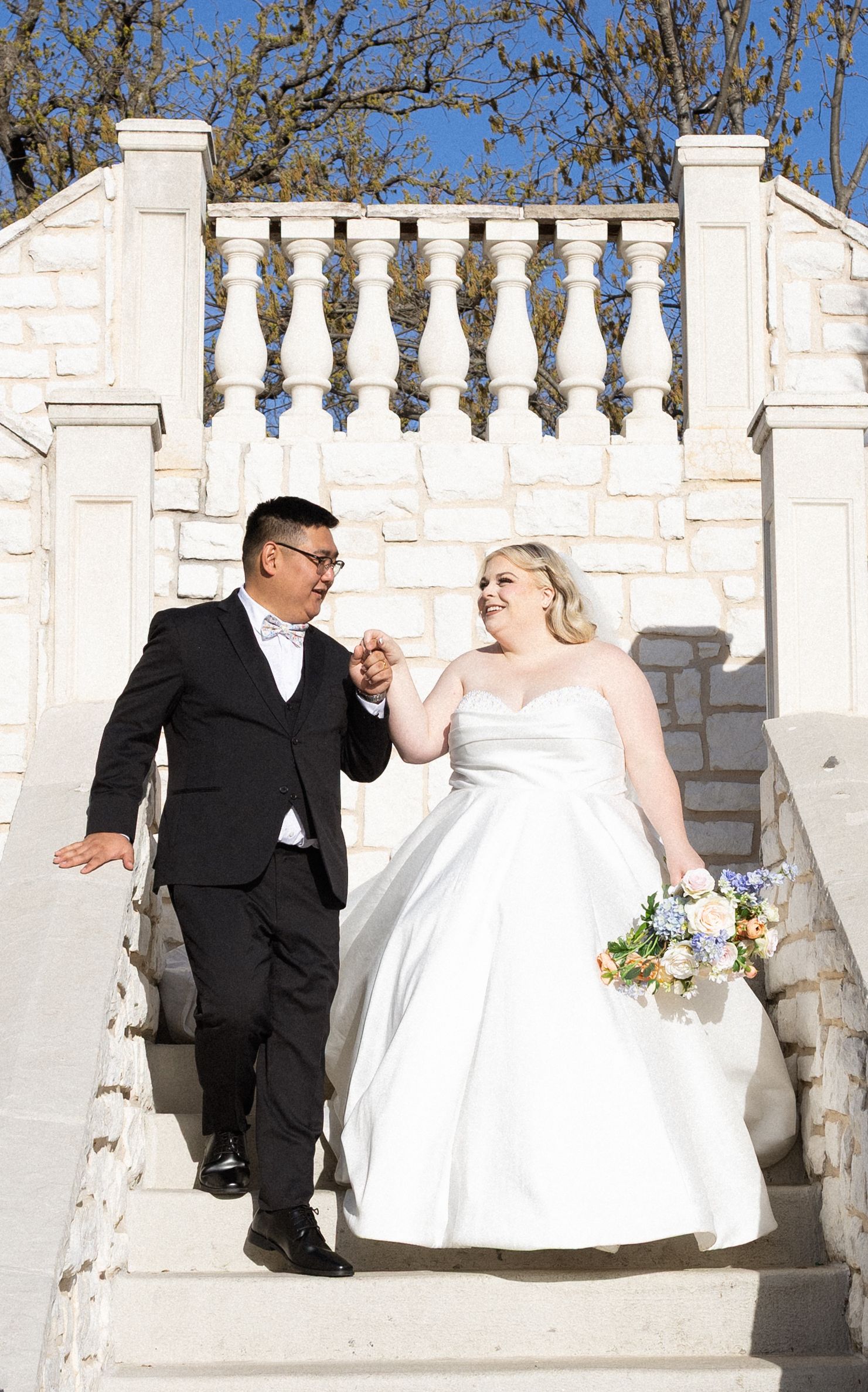 Bride and groom holding hands walking down white stone steps, bride in strapless gown holding bouquet, groom in black tuxedo.