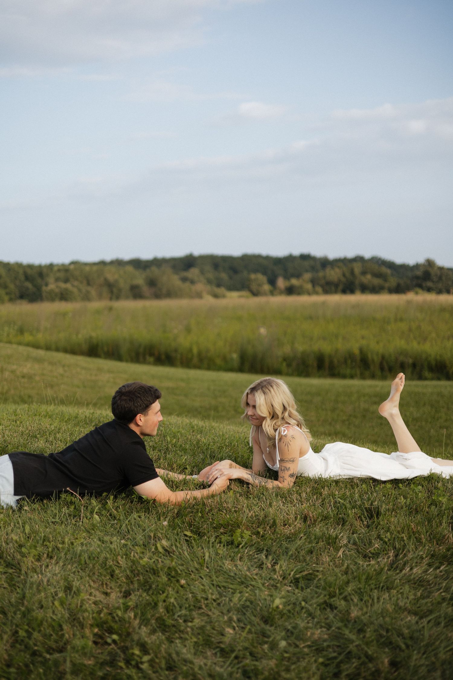 A couple in wedding attire lies on green grass holding hands in a scenic field with trees and blue sky.