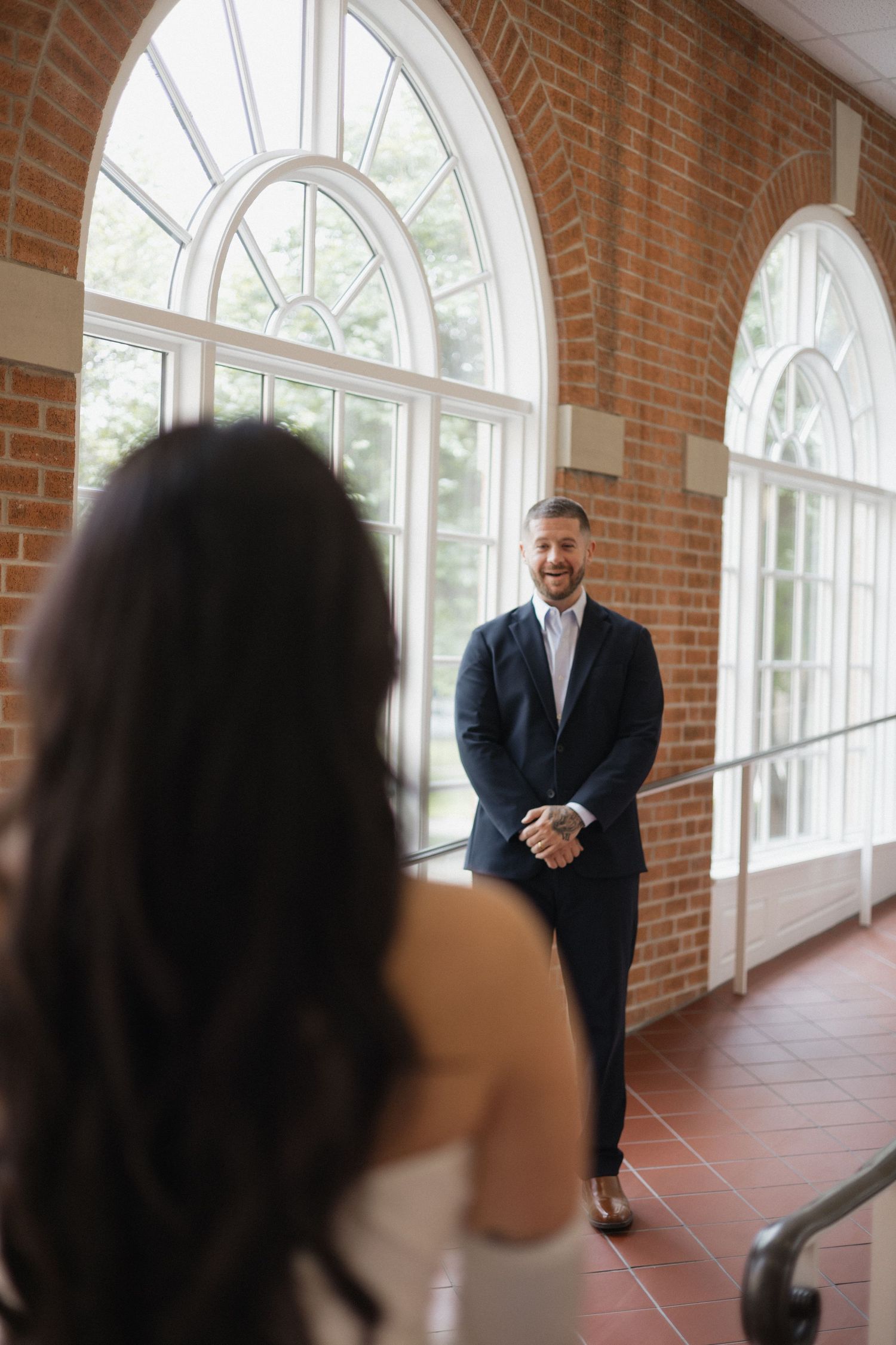 Groom in navy suit smiling at bride during first look moment in elegant brick hallway with arched windows.