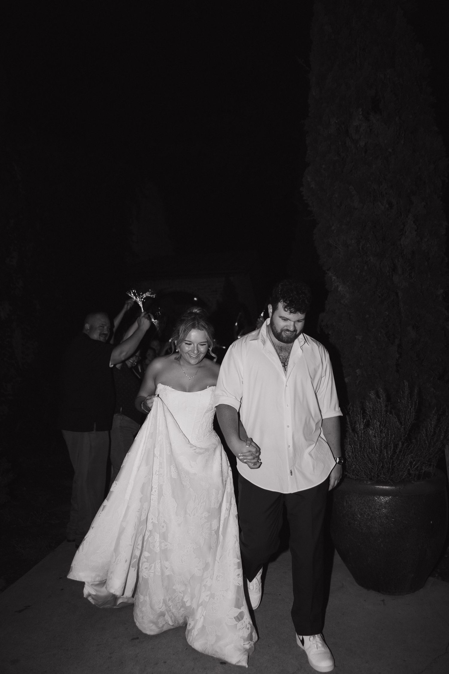 Black and white photo of a bride in a strapless gown and groom in white shirt holding hands at their wedding reception.