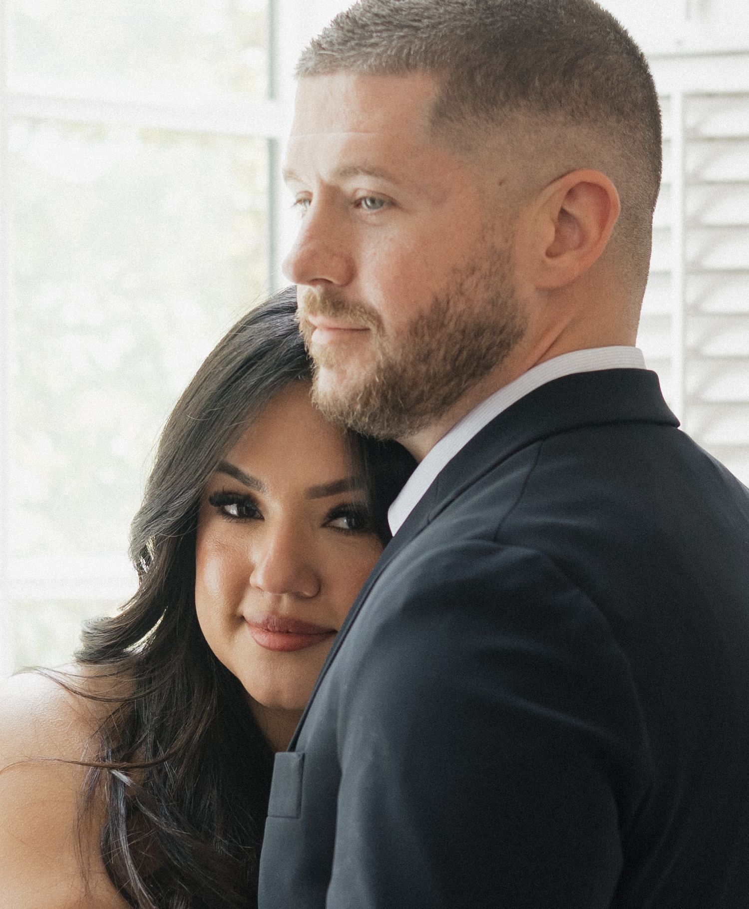 A couple in formal attire poses intimately, the woman leaning against the man in a suit near a bright window.