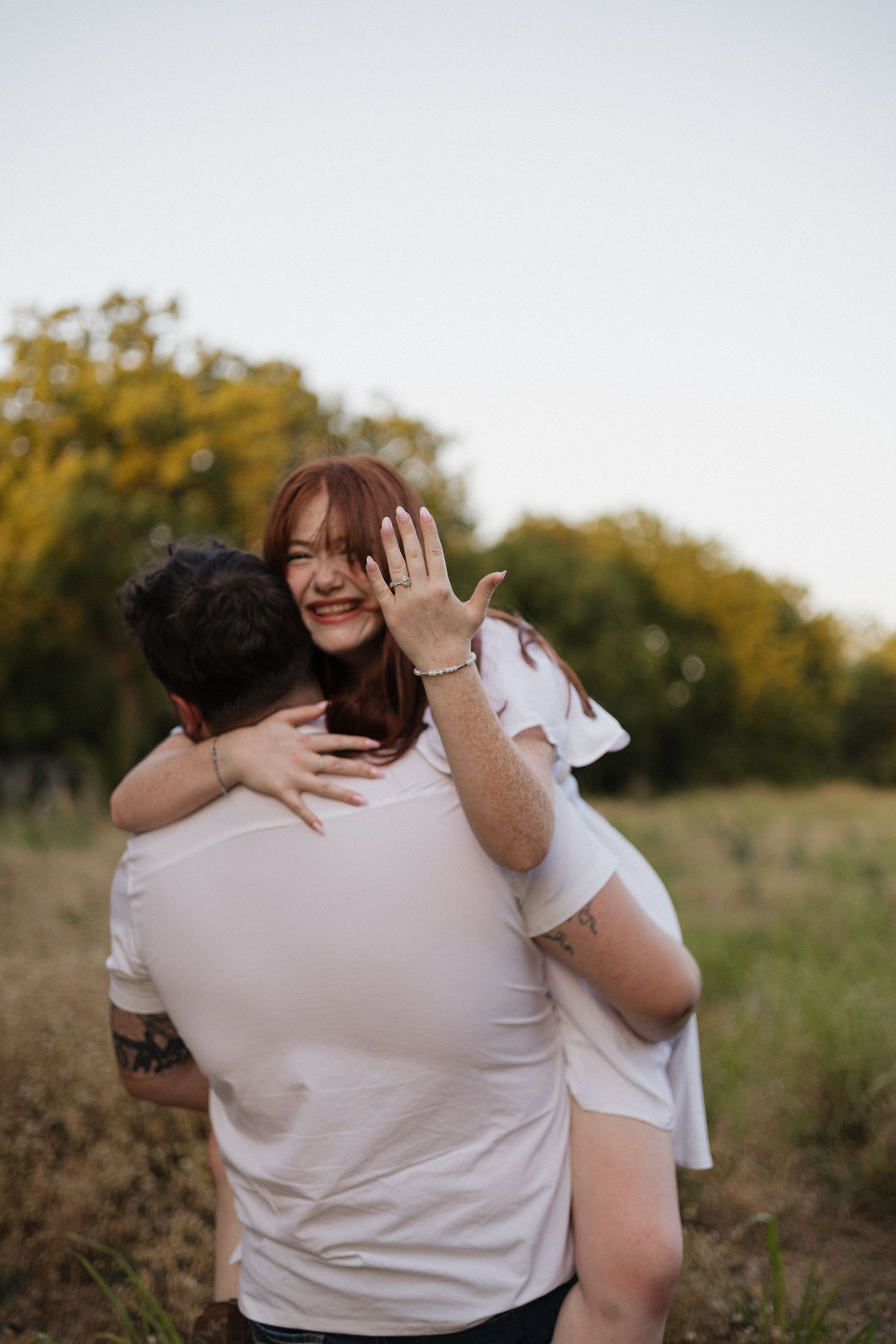 Happy woman showing engagement ring while being lifted by man in white t-shirt in a scenic outdoor field.