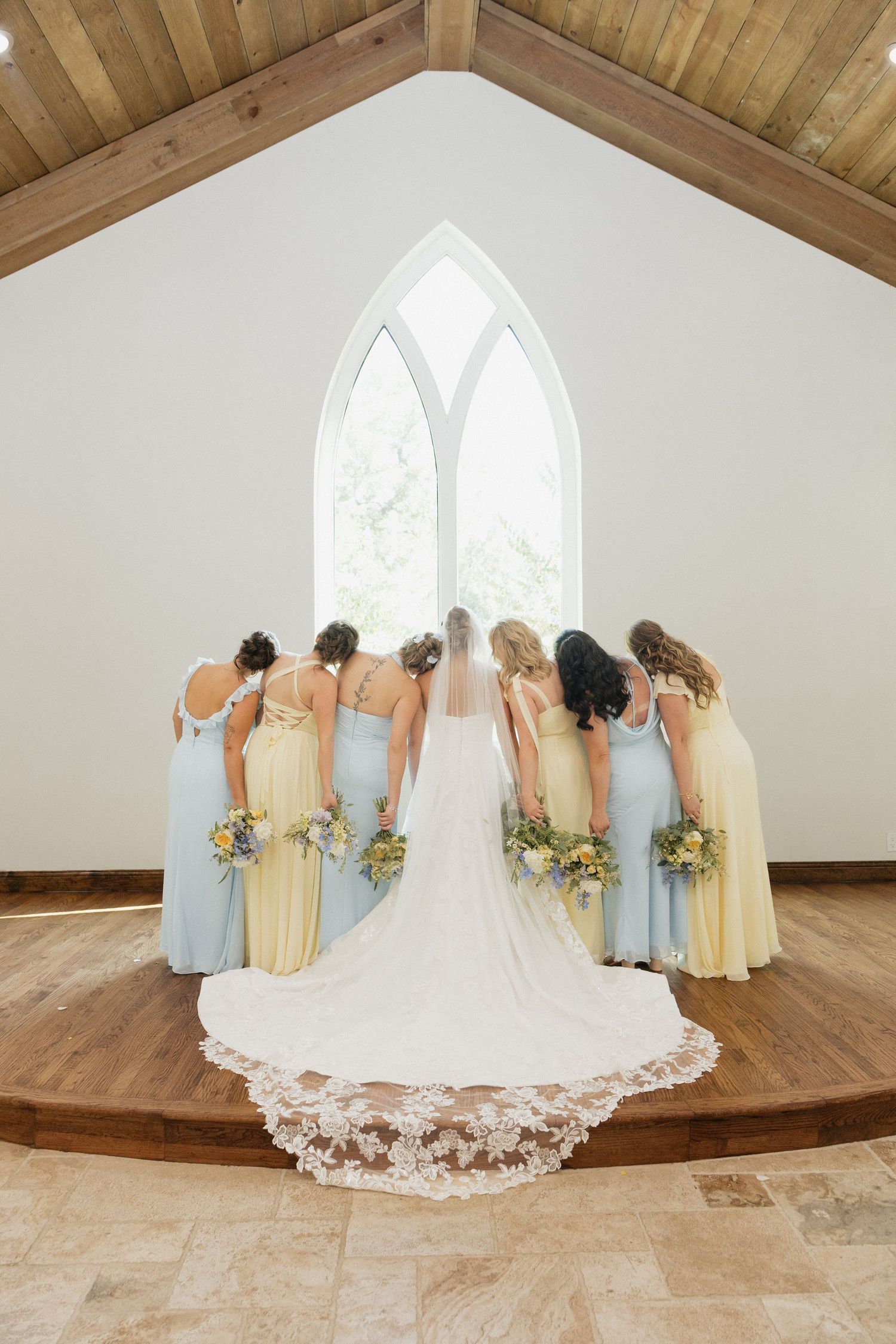 Bride and bridesmaids in pastel blue and yellow gowns stand before a chapel window holding floral bouquets.