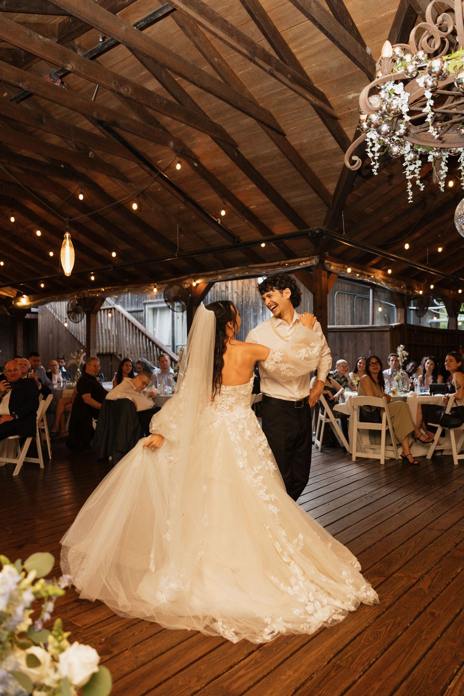 Bride and groom share their first dance in a rustic barn venue with string lights and chandelier. Caddo Mills Texas wedding photographer