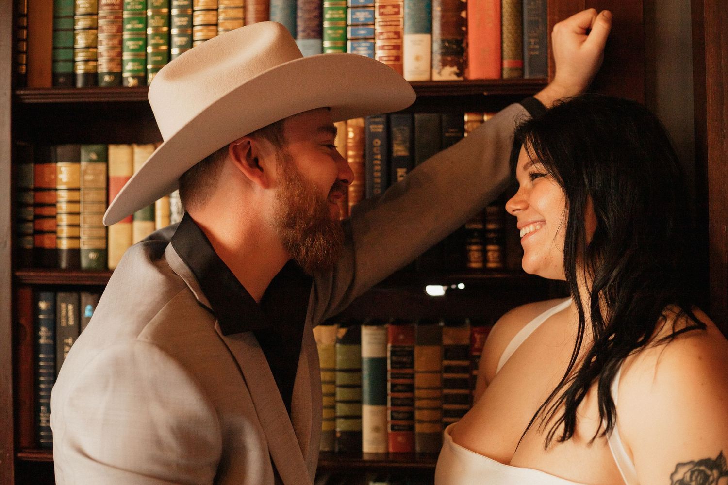 Intimate couple portrait session with cowboy hat against library bookshelf backdrop.