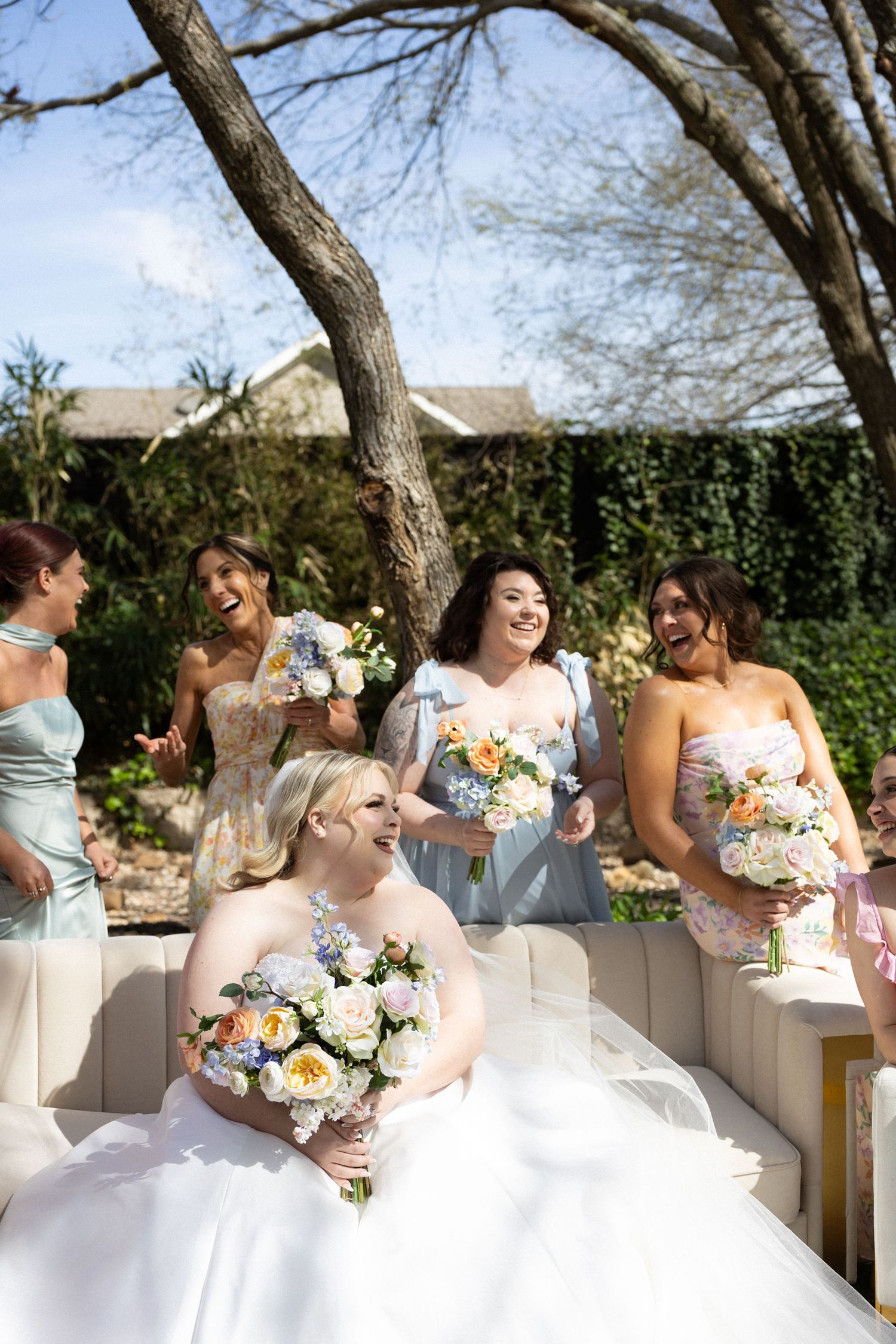 Bride in white gown holds floral bouquet surrounded by laughing bridesmaids in blue dresses at outdoor wedding.
