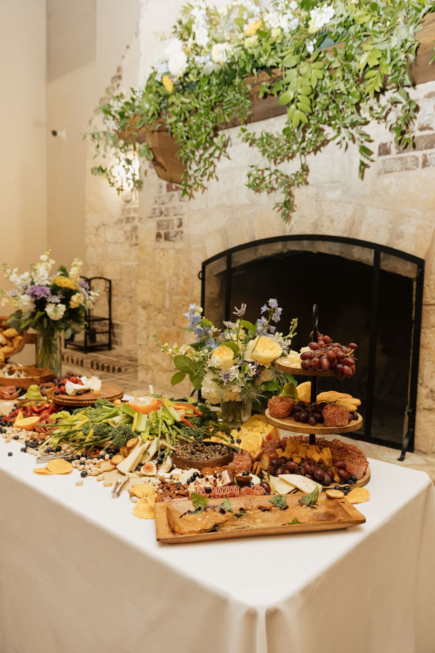 Elegant grazing table with charcuterie, fruits, and florals displayed in front of a stone fireplace with greenery.