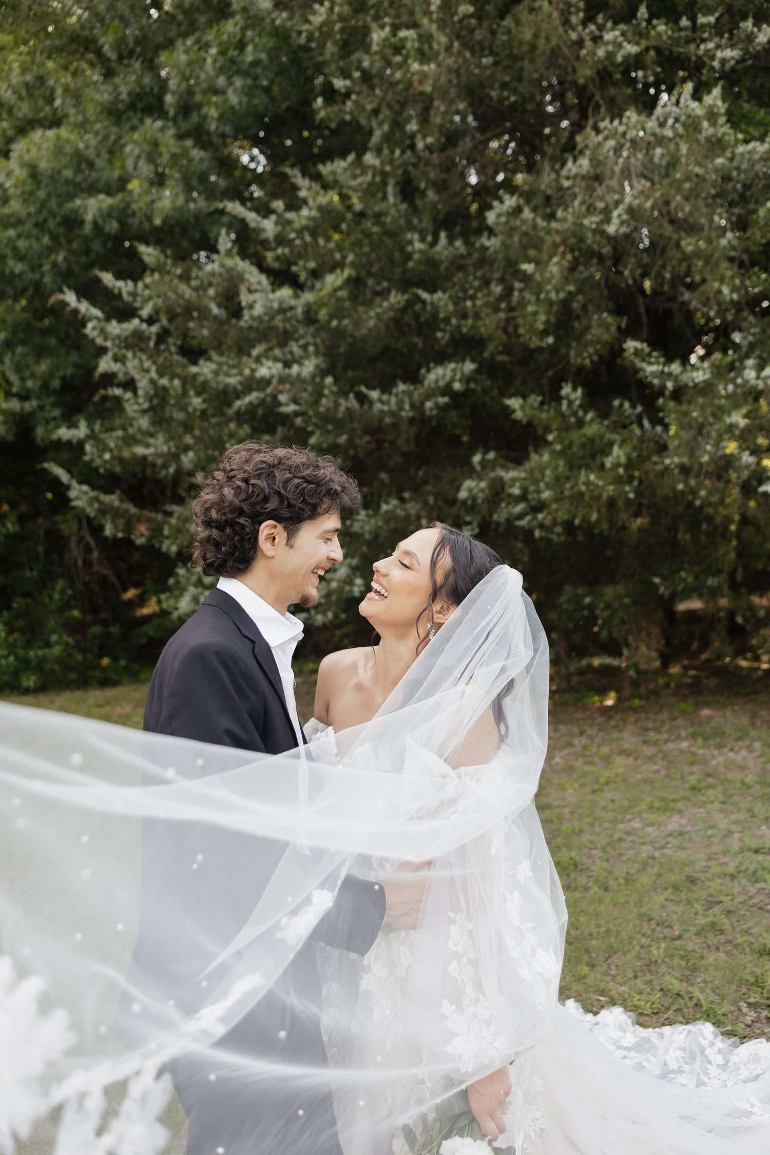 Bride and groom share a romantic moment under a flowing veil with lush green trees in the background.