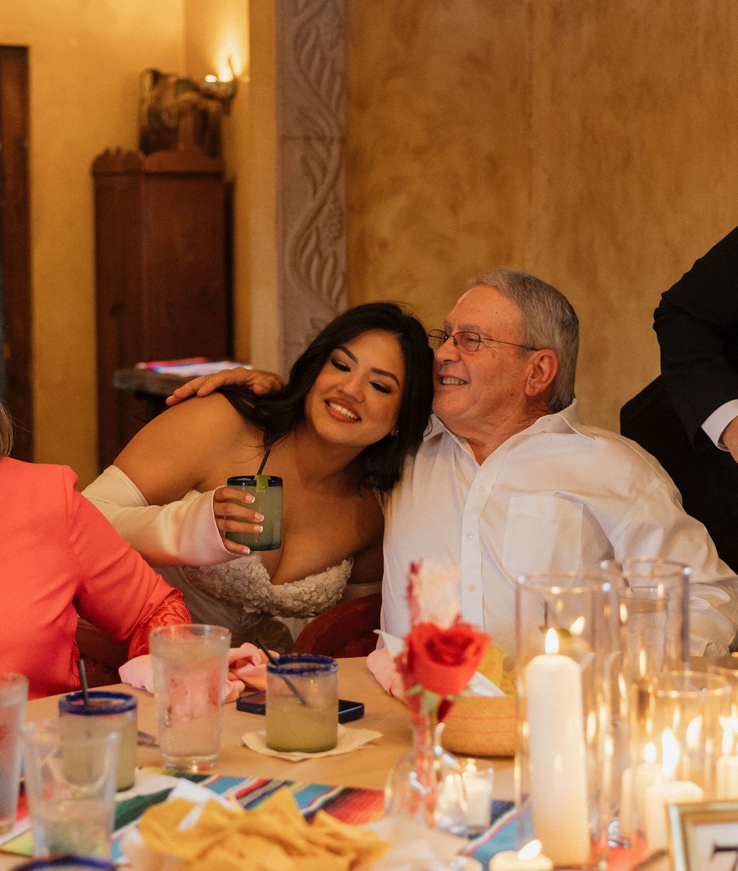 A bride and elderly man smile warmly at a candlelit wedding reception dinner table with colorful decor.