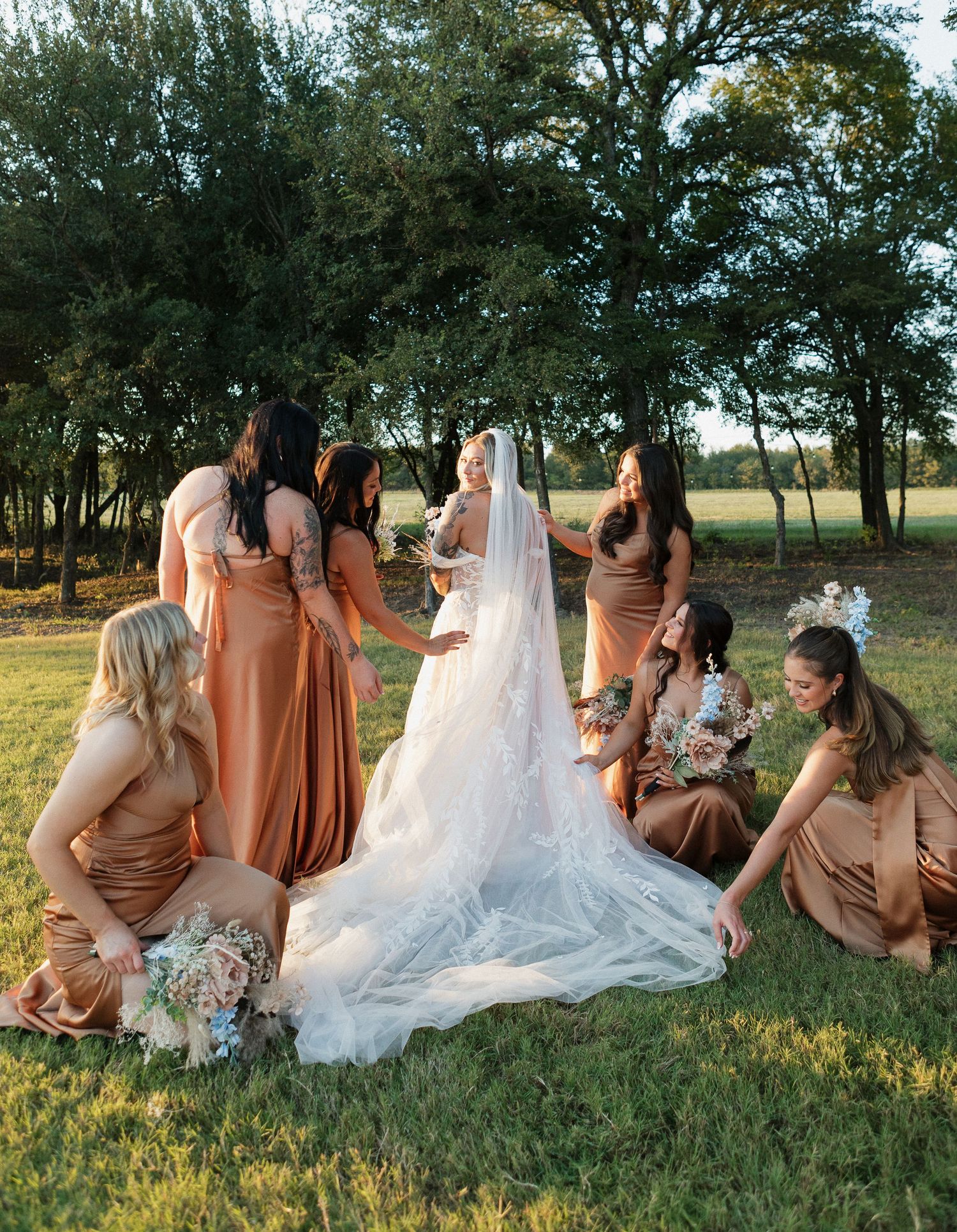 Bridesmaids in peach dresses sitting with bride on grassy field at sunset.