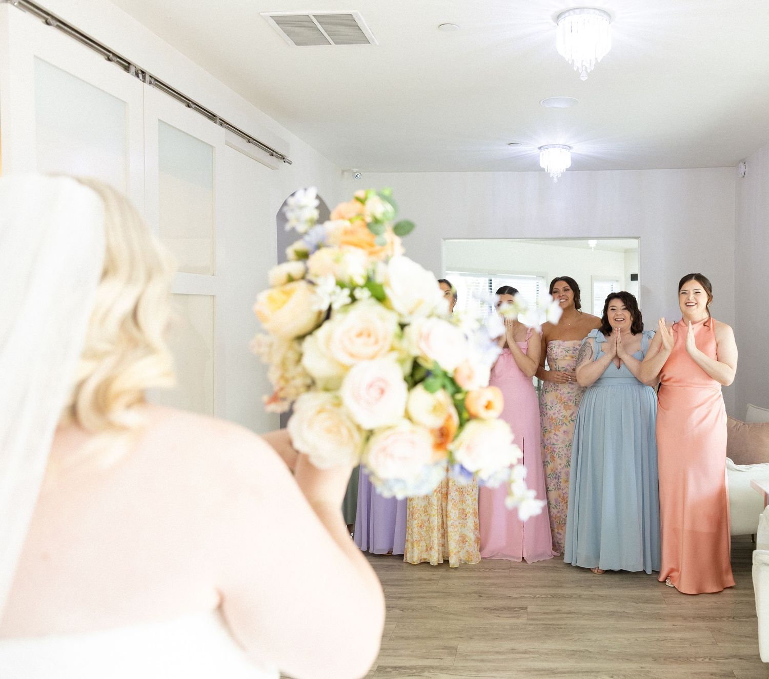 Bridesmaids in pastel gowns react with joy as bride in white wedding dress holds bouquet of peach and white roses.