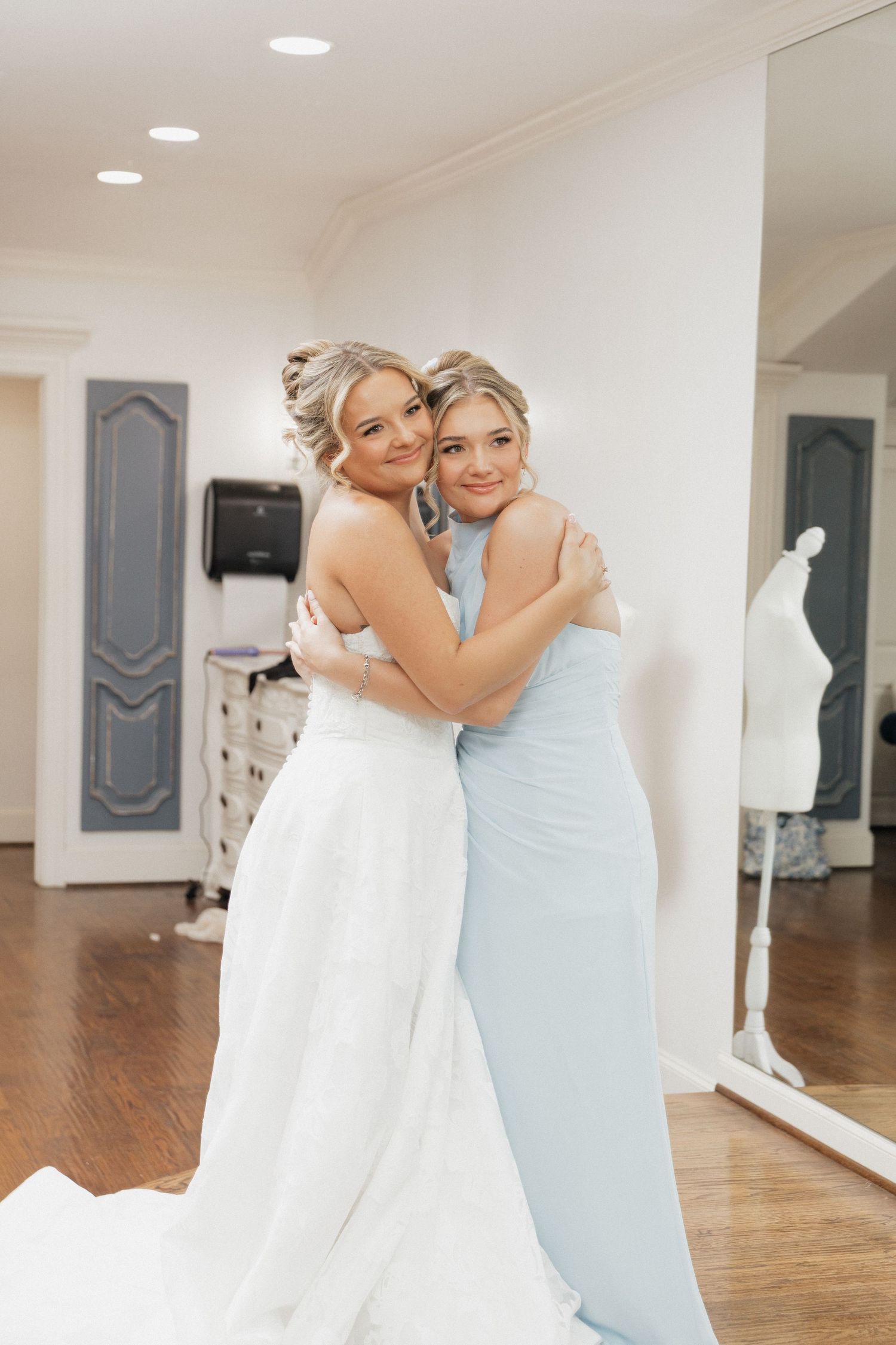Bride in white gown hugging bridesmaid in light blue dress, smiling in elegant bridal suite.