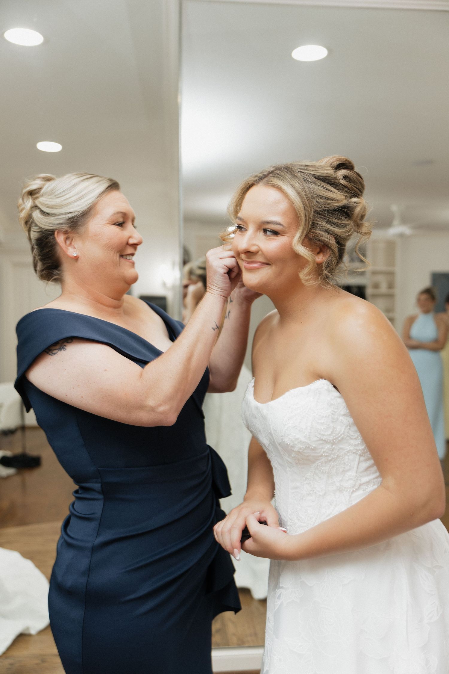 A mother in a navy dress helps her daughter put on earrings as the bride prepares in her white strapless wedding gown.
