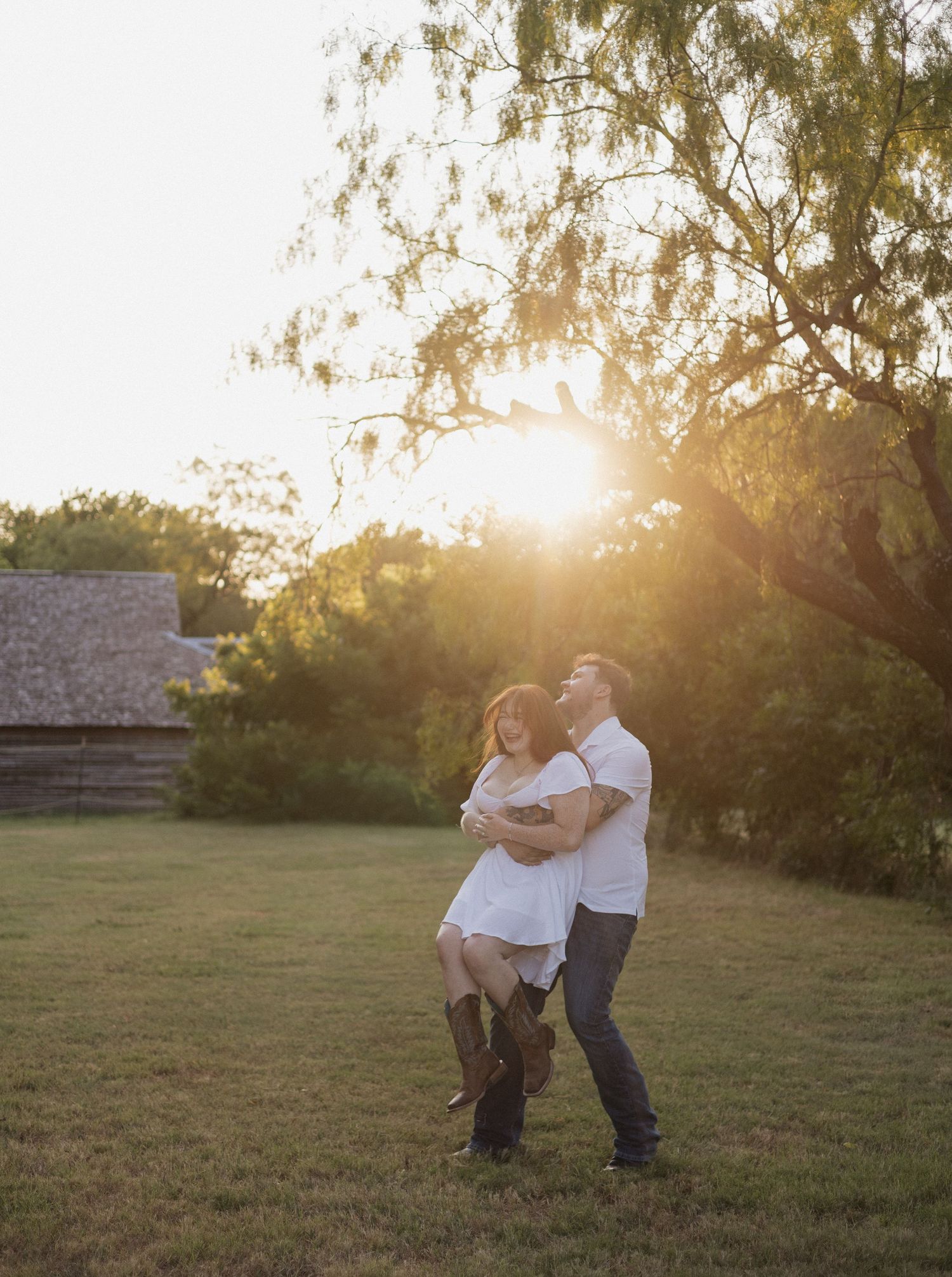 Couple dancing in golden sunset light on green grass field near rustic barn and trees.