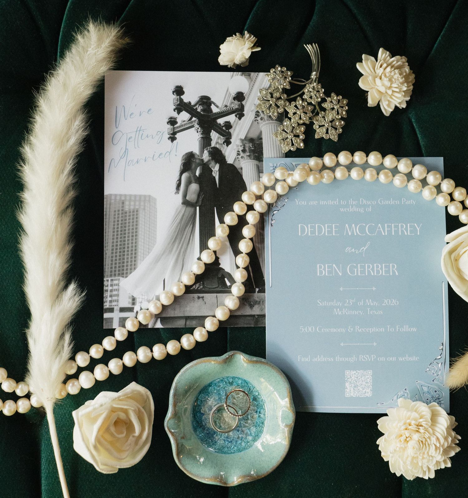 Elegant wedding invitation suite with blue stationery, pearl beads, white flowers, and pampas grass on green velvet.