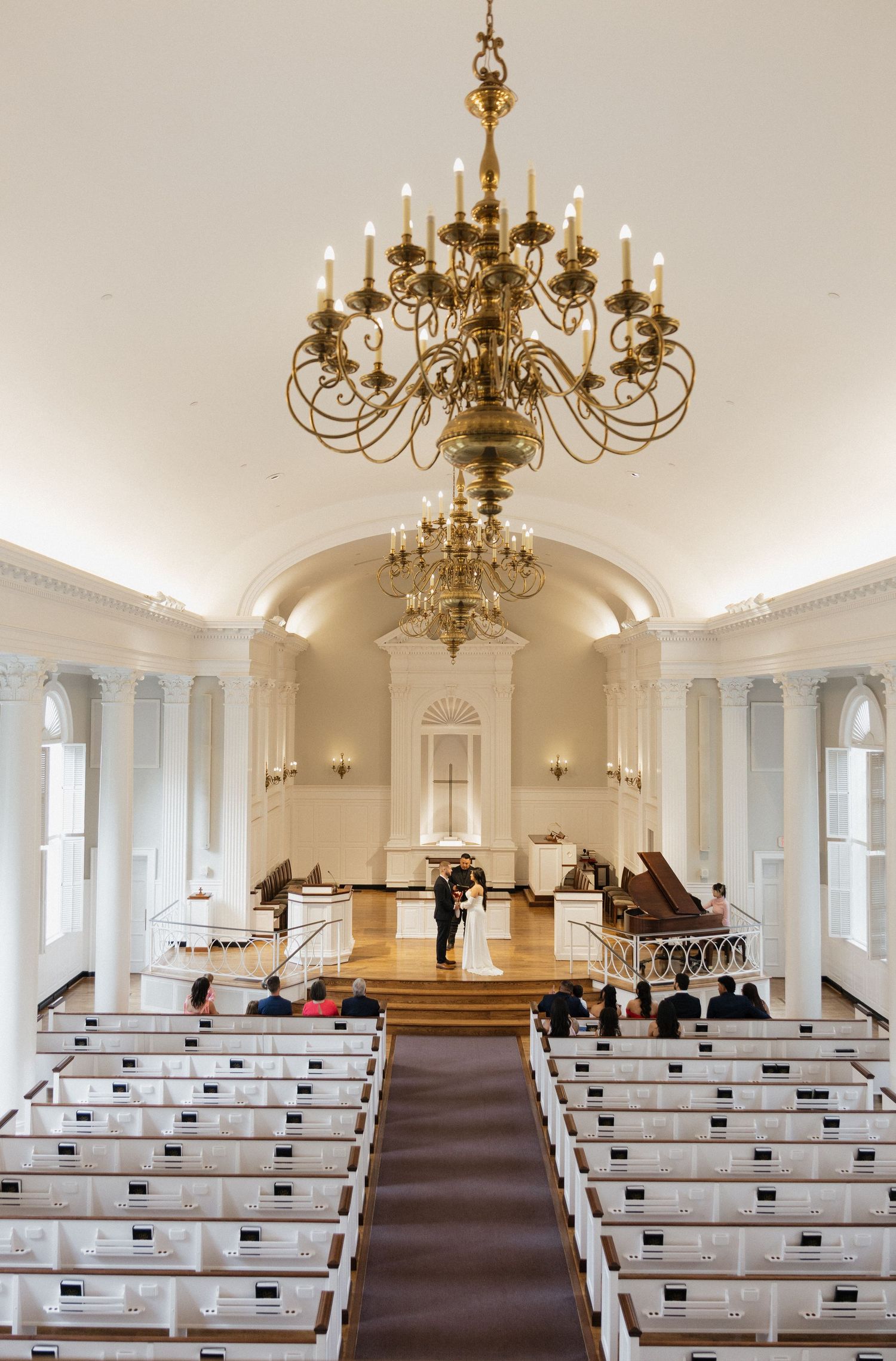 Elegant church wedding ceremony with gold chandelier, white pews, and couple at altar in classic chapel.