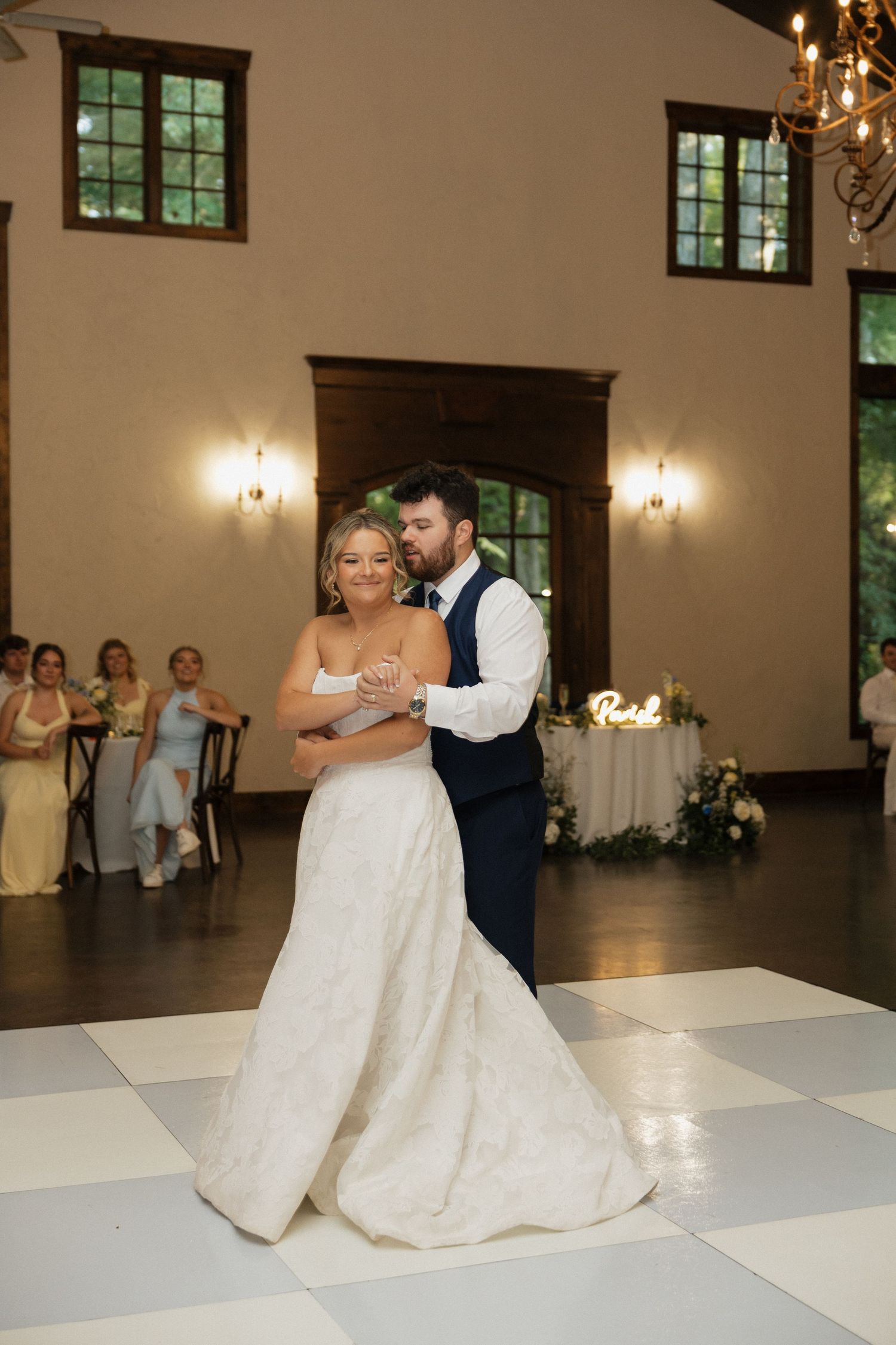 Newlyweds share romantic first dance on white checkered floor in elegant warmly lit reception hall.