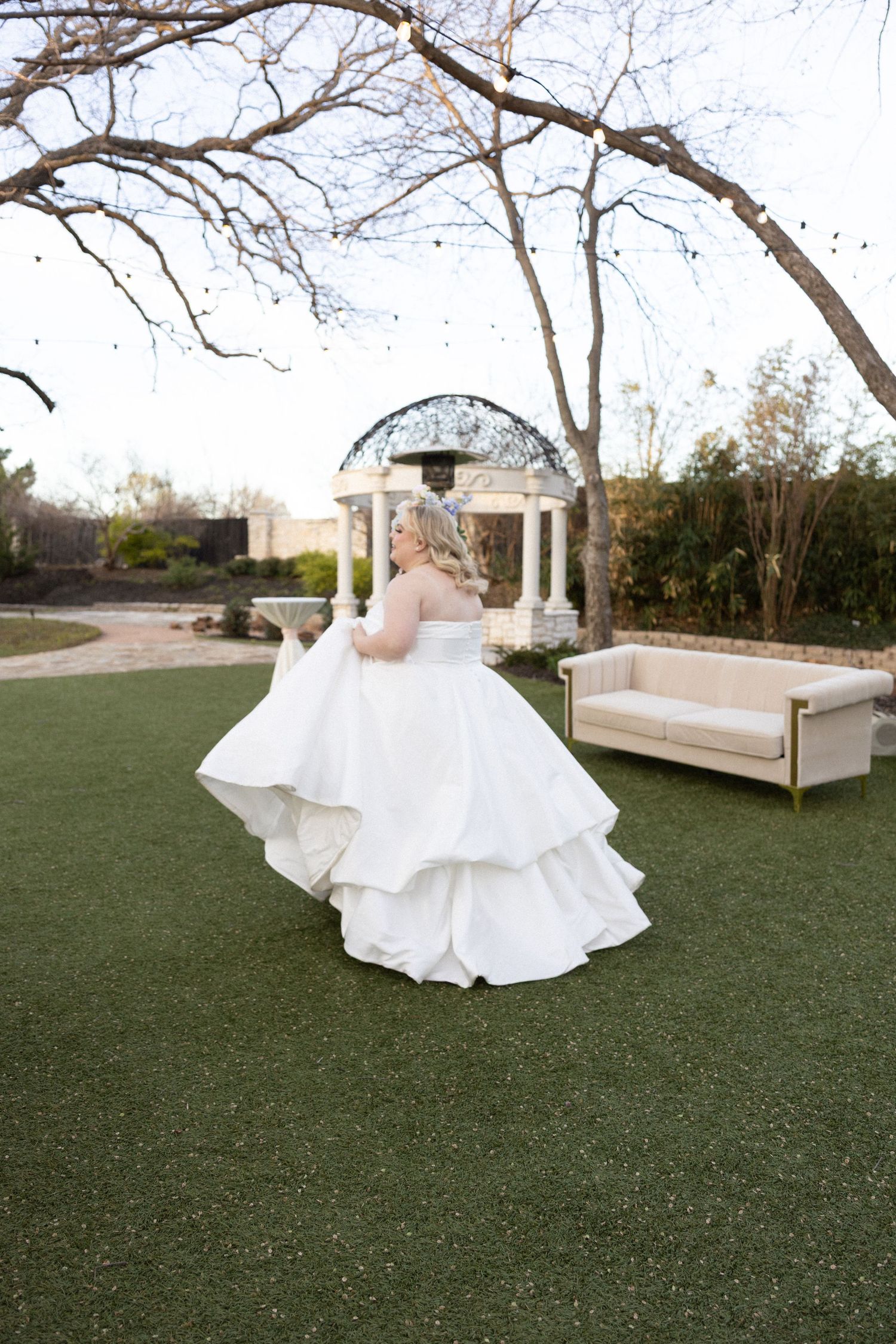 Bride twirls in a layered white ballgown on green lawn with gazebo backdrop at outdoor wedding venue.