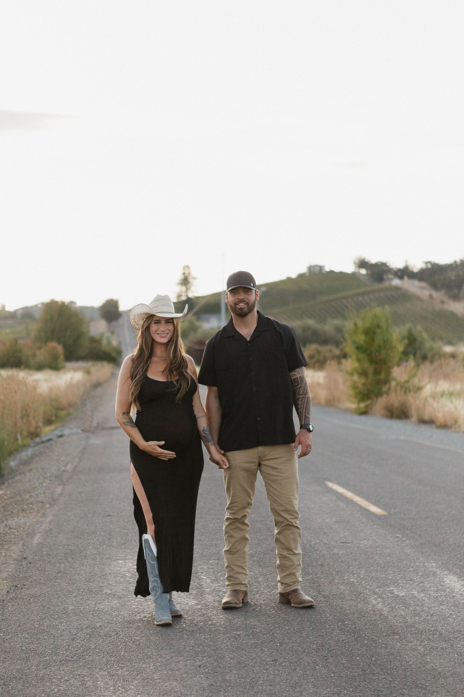 A couple in black and khaki outfits walks together on an empty road surrounded by desert landscape at sunset.