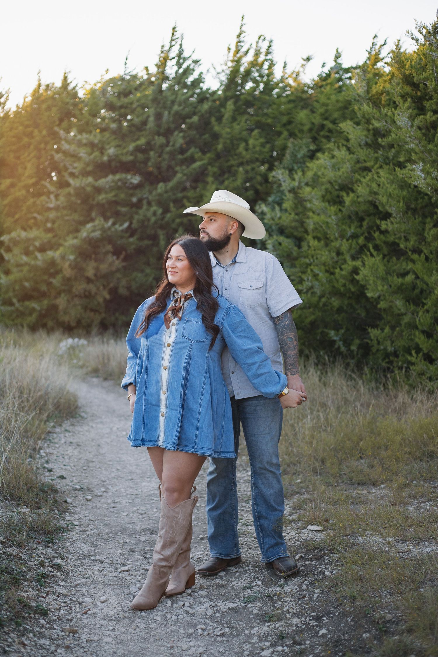 Couple in denim outfits standing on dirt path with evergreen trees in background.