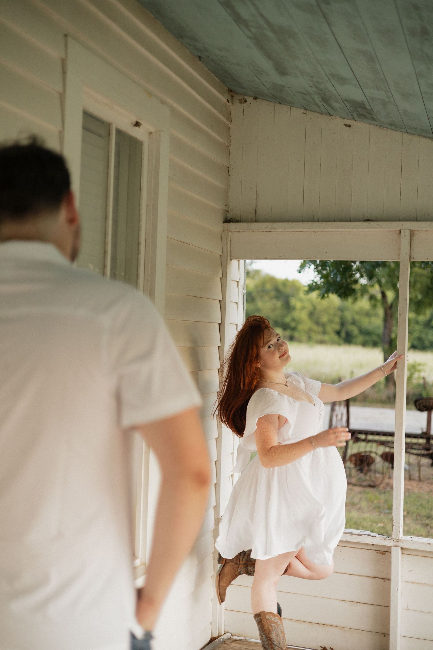 A woman in a white dress and cowboy boots leans against an open window on a rustic porch while a man in white approaches.