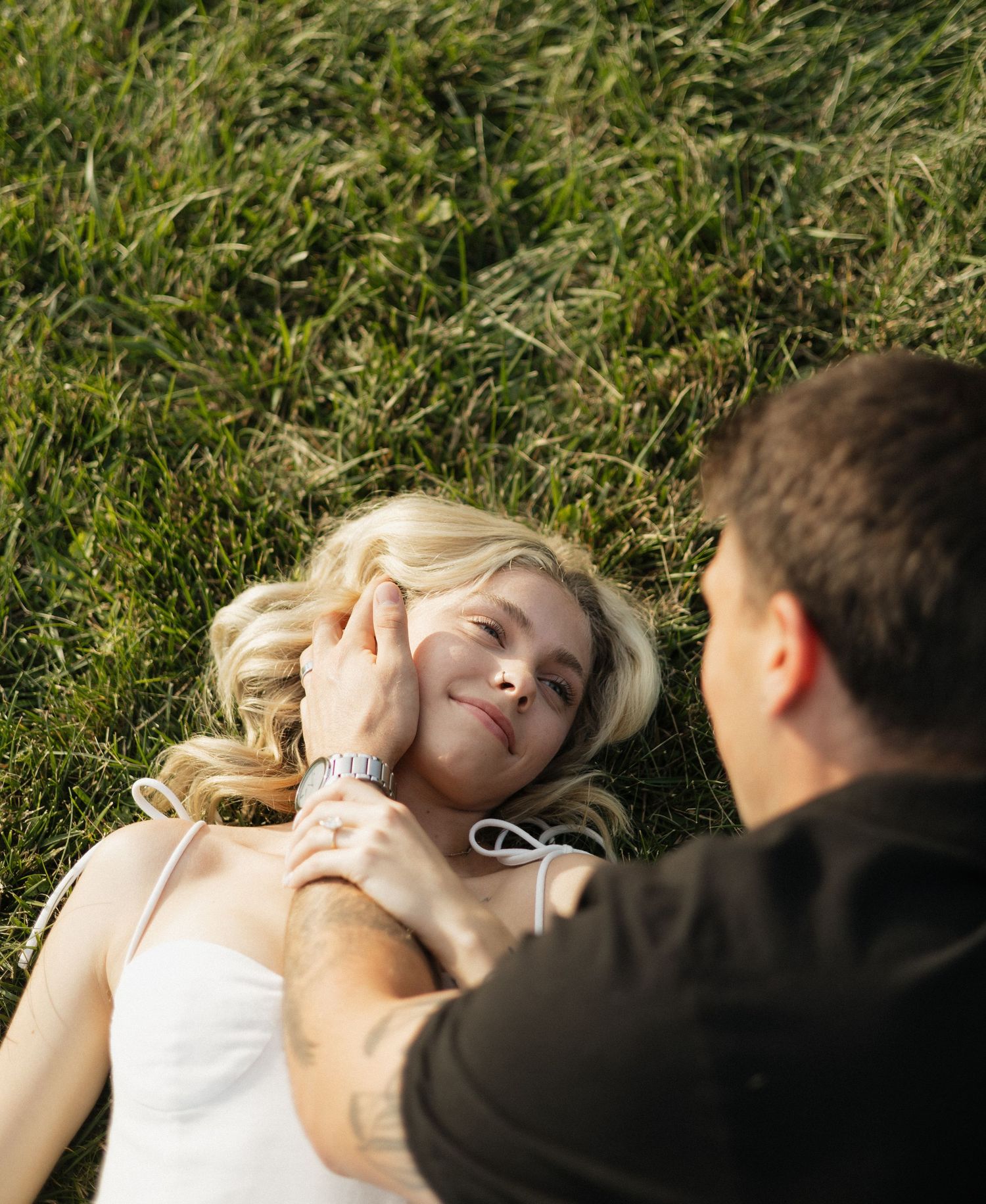 Couple relaxing in green grass, man gently touching blonde woman's face while she smiles up at him.